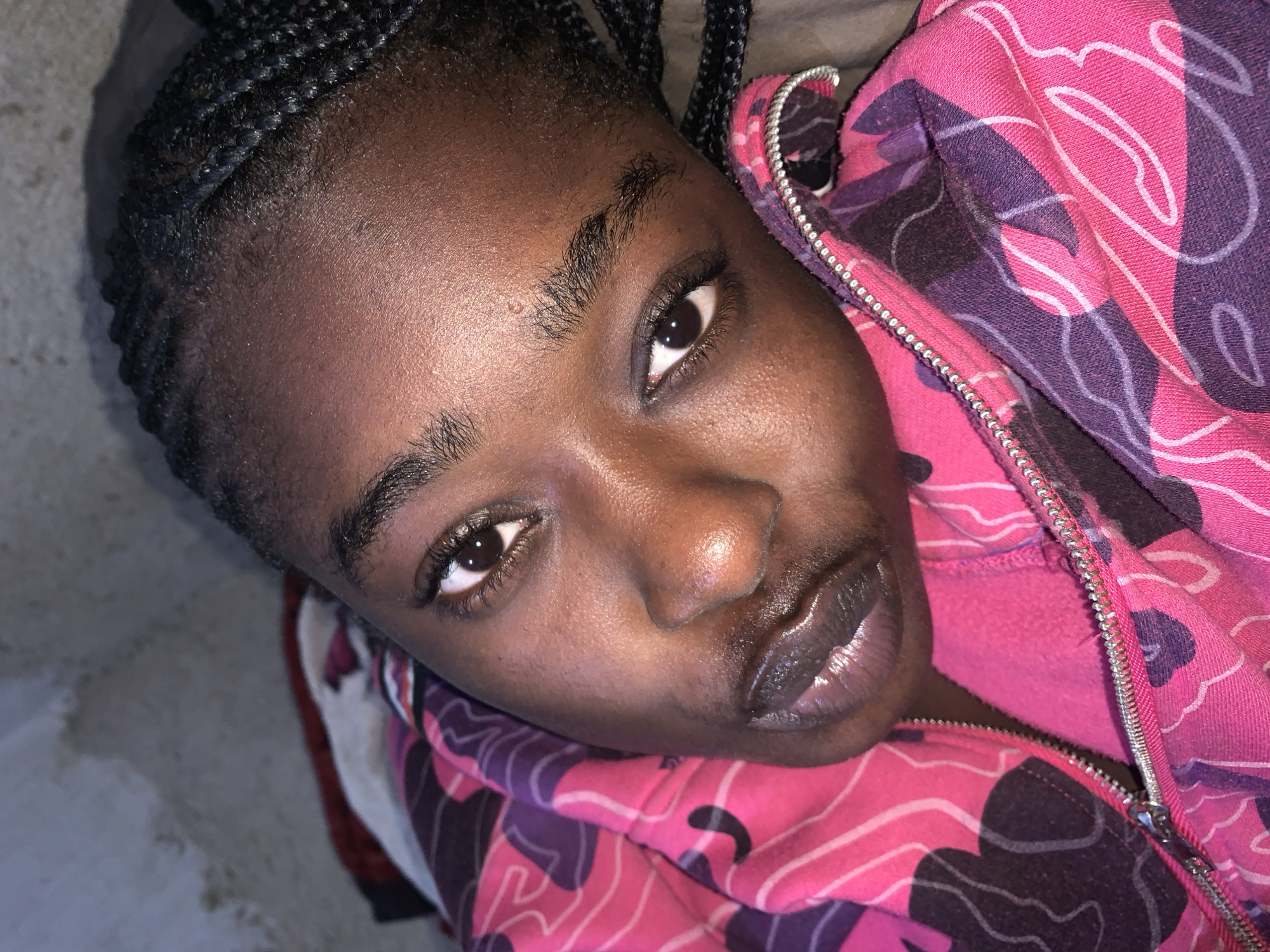 The image is a close-up, overhead shot of a young Black woman with braided hair, wearing a pink and purple camouflage-patterned hooded sweatshirt. She is lying down, with her head resting on her shoulder, and her gaze is directed towards the camera. Her skin is dark and slightly shiny. Her lips are full and appear to be glossed. The background is blurred and out of focus, showing a mottled grey surface. The setting appears to be indoors. There is no specific information in the image to indicate the location is in Rafin Par, Nigeria.