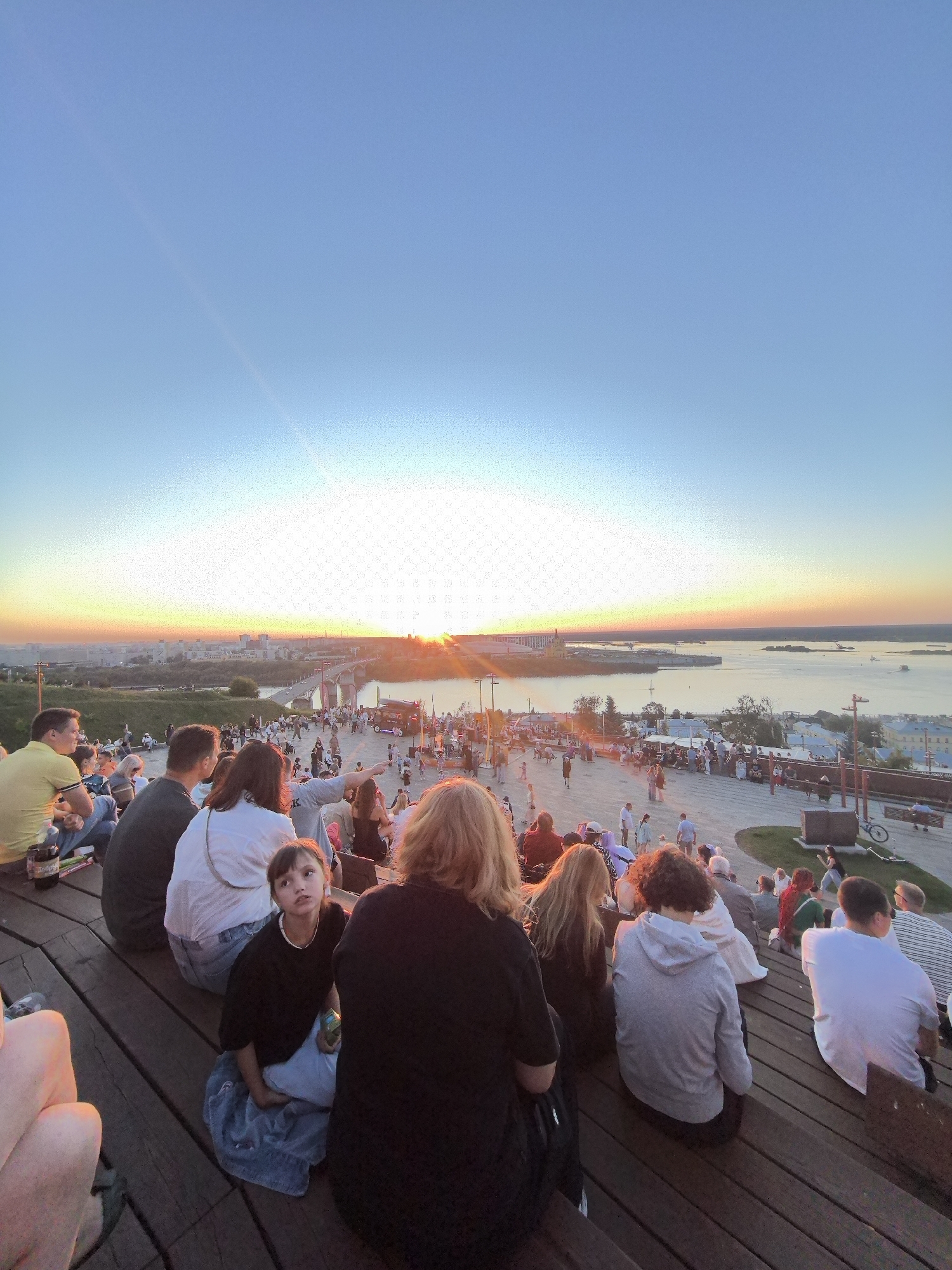 The image shows a crowd of people sitting on wooden bleachers, looking out at a sunset over a city skyline and a river. The sun is setting on the horizon, casting a warm glow over the scene. A bridge spans the river in the distance, and boats can be seen on the water. The city appears to be Nizhny Novgorod, Russia, based on the distinctive architecture and the Kremlin visible on the hill. The people are casually dressed, and some are pointing towards the sunset or talking to each other. The atmosphere seems relaxed and festive, with a sense of community gathering to enjoy the natural beauty of the sunset. The foreground is dominated by the wooden bleachers and the backs of several people seated on them.