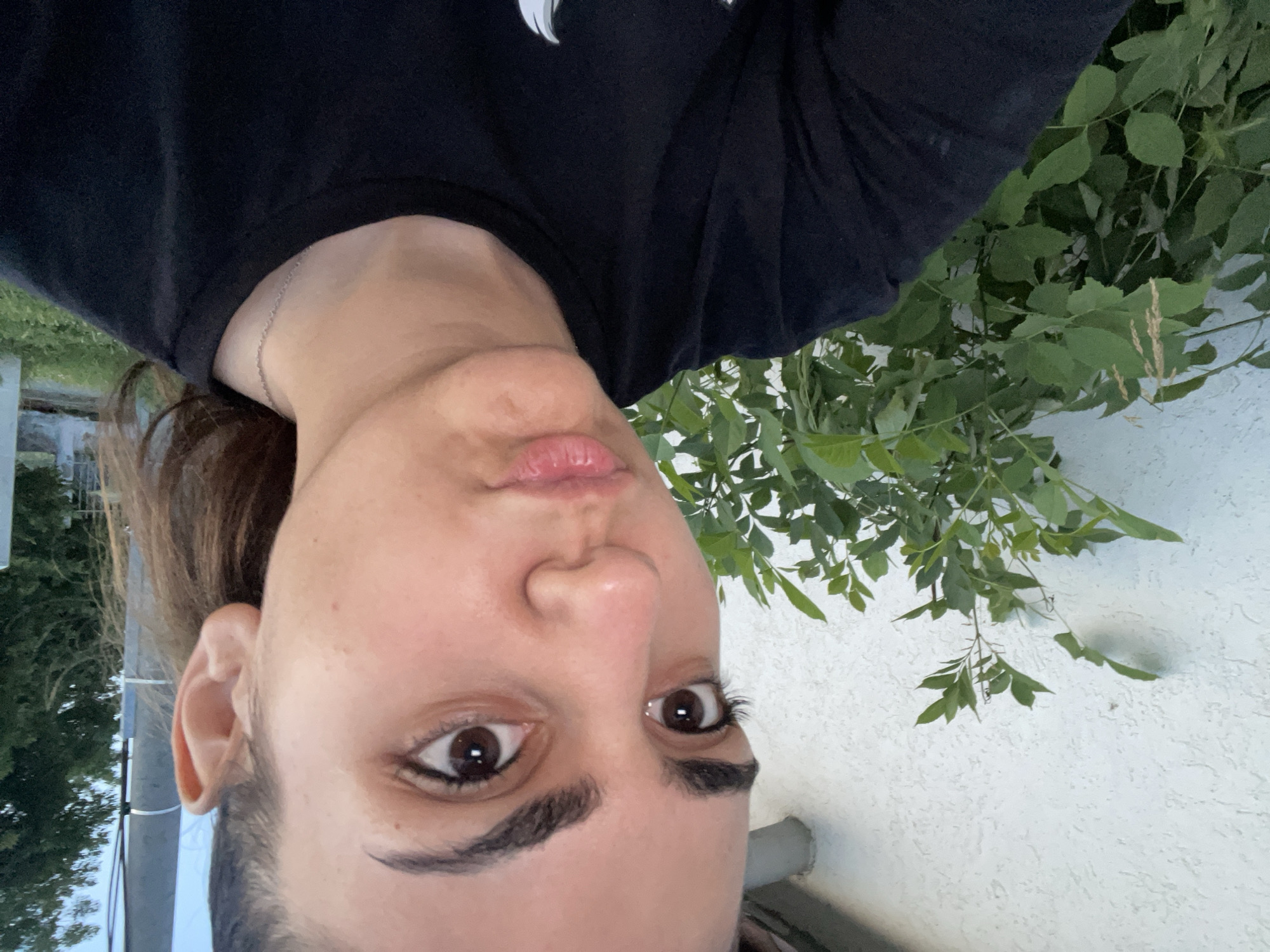 A medium-close up outdoor portrait captures a young adult female in Debelt, Bulgaria. She has dark brown hair, pulled back, and her dark brown eyes are wide open, directed towards the viewer. Her lips are pursed in a pouting expression. She is wearing a black short-sleeved t-shirt and a thin silver chain necklace around her neck.
To the subject's left, a dense green leafy plant, possibly a bush or small tree, grows against a white, stucco-textured building wall. Some lighter, dried, reedy stalks are visible among the green leaves. A small, light-colored, circular object, appearing to be a vent or pipe opening, is affixed to the upper right section of the visible wall.
To the subject's right, a light gray concrete utility pole stands vertically, partially obscured by her head and shoulder. Beyond the pole, dense green tree foliage under a pale blue sky completes the background, indicating an outdoor, possibly semi-urban or residential, setting.