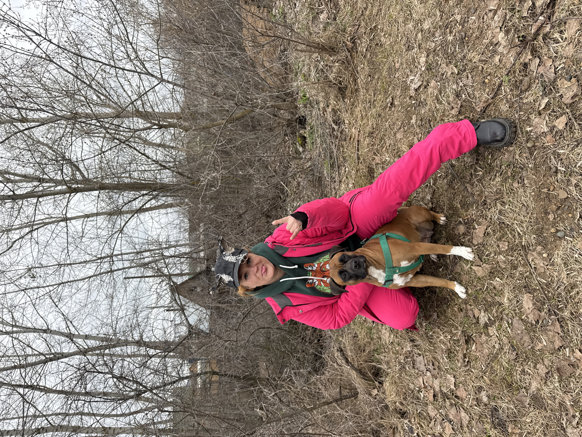 A person and a dog are captured in an outdoor portrait, kneeling on dry, grassy ground with a background of bare trees. The image is oriented vertically (rotated 90 degrees clockwise from how it was likely taken).
The primary subject is a person, likely female, wearing a vibrant pink jacket and matching pants, a dark green hoodie with an orange and white animal face graphic on the front, and a dark, distressed cap adorned with metal studs or spikes. She is looking directly at the camera with her tongue slightly extended and one hand raised in a gesture, possibly a "rock on" or peace sign. She wears black boots on her feet.
Beside her, a brown and white Boxer dog lies calmly on the ground, also looking towards the camera. The dog has a fawn-colored coat with white markings on its chest, muzzle, and paws, and is wearing a green harness.
The setting appears to be a wooded area or forest during late winter or early spring in Chaikovsky, Russia. The ground is covered with dried brown leaves and sparse, dead grass, indicating the dormant season. The background is filled with numerous deciduous trees and shrubs, all bare of leaves, their intricate branches creating a delicate, skeletal pattern against an overcast, light grey sky. The overall light suggests a cloudy day. No explicit text is visible in the image.