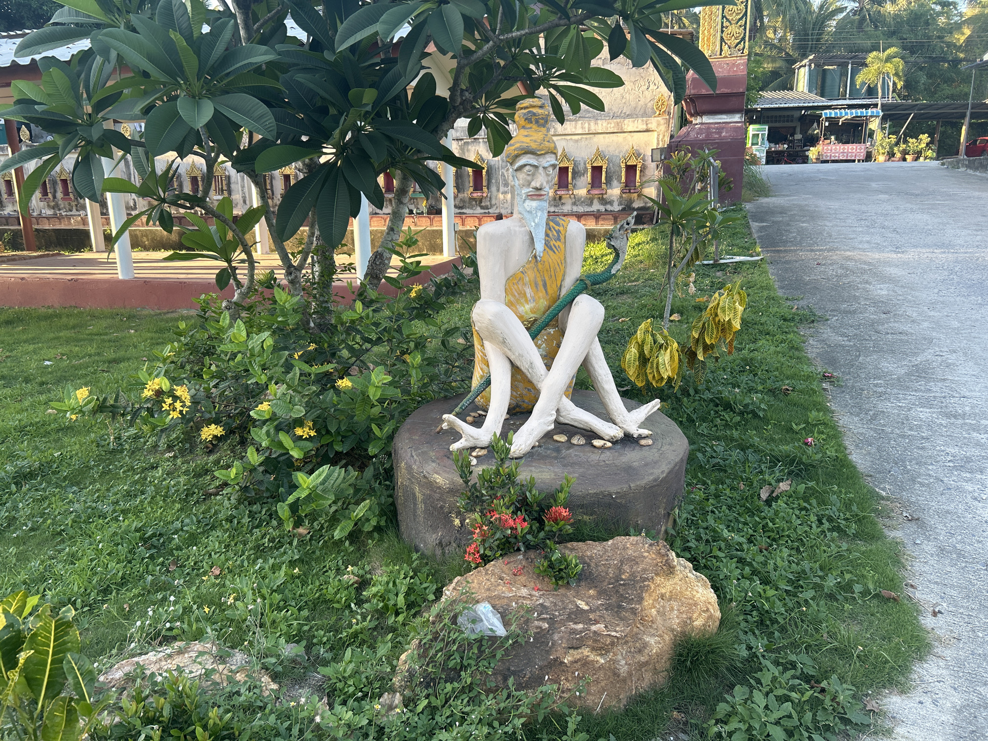 The image captures a statue of an elderly man with a white beard and a yellow robe, seated on a large rock. The statue is surrounded by lush green grass, small yellow flowers, and a leafy green tree. In the background, a weathered building with intricate yellow and red details, possibly a temple, stands tall. A paved pathway runs along the right side of the image, leading to a covered structure with tables and chairs, suggesting a public space or market. The overall setting appears to be outdoors, in a tropical climate, given the dense vegetation and the style of the architecture. The scene is bathed in natural light, indicating it was taken during the daytime.