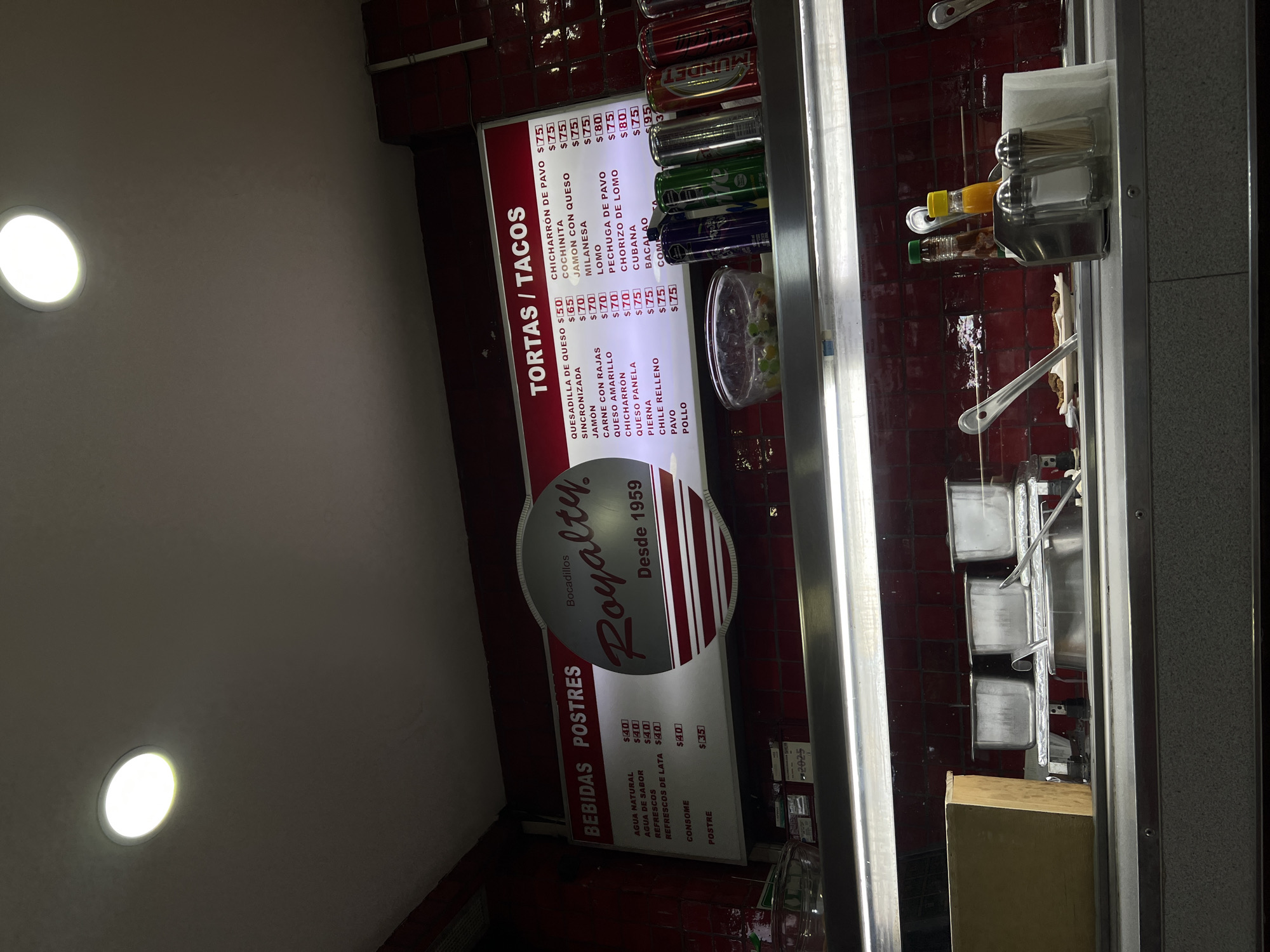 The image shows the interior of a small food establishment, most likely a "torteria" or "taqueria" in Mexico City, Mexico. A vertically oriented menu board is prominently displayed, listing various "tortas" (sandwiches) and "tacos" with their prices, along with "bebidas" (drinks) and "postres" (desserts). The logo "Royalty. Bocadillos. Desde 1959" is visible above the menu items. Canned drinks, condiments, and napkins are arranged on a shelf. The red tiled wall indicates a traditional Mexican environment. The lighting is provided by recessed ceiling lights.