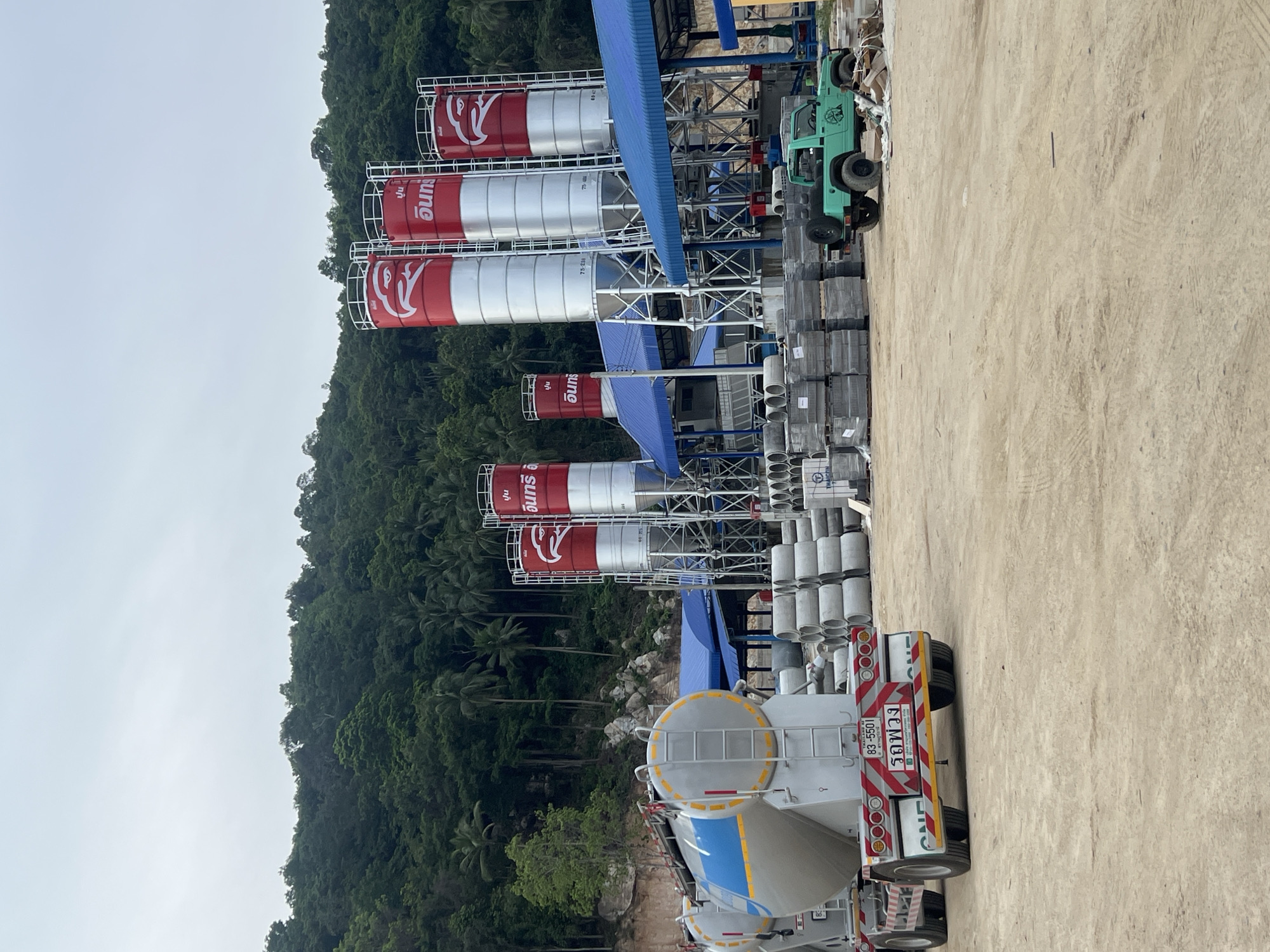 The image depicts an industrial scene in ตำบลเกาะพะงัน, Thailand, with a focus on a cement factory or construction material storage area. In the foreground, a large cement truck with a cylindrical tank is parked, facing left. The truck has a license plate that reads "83-5501" and text in Thai that appears to be "รถพ่วง" (rot phwong), meaning "trailer truck." To the right of the truck, a stack of concrete pipes and other construction materials are visible, along with a small, bright green off-road vehicle.
Dominating the midground and background are several tall, industrial silos, painted red and silver, each adorned with a white eagle logo. These silos are part of a structure with blue roofing. Behind the industrial site, a lush, densely forested hillside rises, characteristic of the tropical environment of Koh Phangan. The sky is overcast, suggesting a cloudy or hazy day. The ground is a light brown, dusty earth, indicating an active construction or industrial zone. The overall impression is one of a functioning industrial facility within a natural, verdant setting.