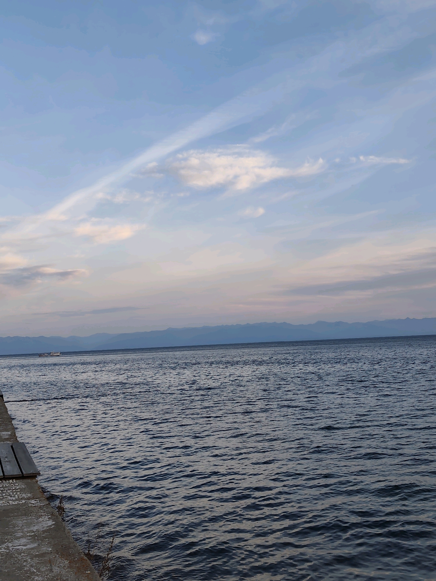 The image depicts a serene lake scene, likely taken from a pier in Listvyanka, Russia, on the shores of Lake Baikal. The foreground features the edge of a concrete pier with wooden planks. The water is a deep blue with small ripples, suggesting a calm day. In the distance, a hazy mountain range forms the horizon, softened by the atmospheric perspective. The sky is a blend of soft blues and pinks, with wispy white clouds stretching across it. A small white boat is visible on the water, adding a touch of scale to the vastness of the lake. The overall impression is one of tranquility and natural beauty.