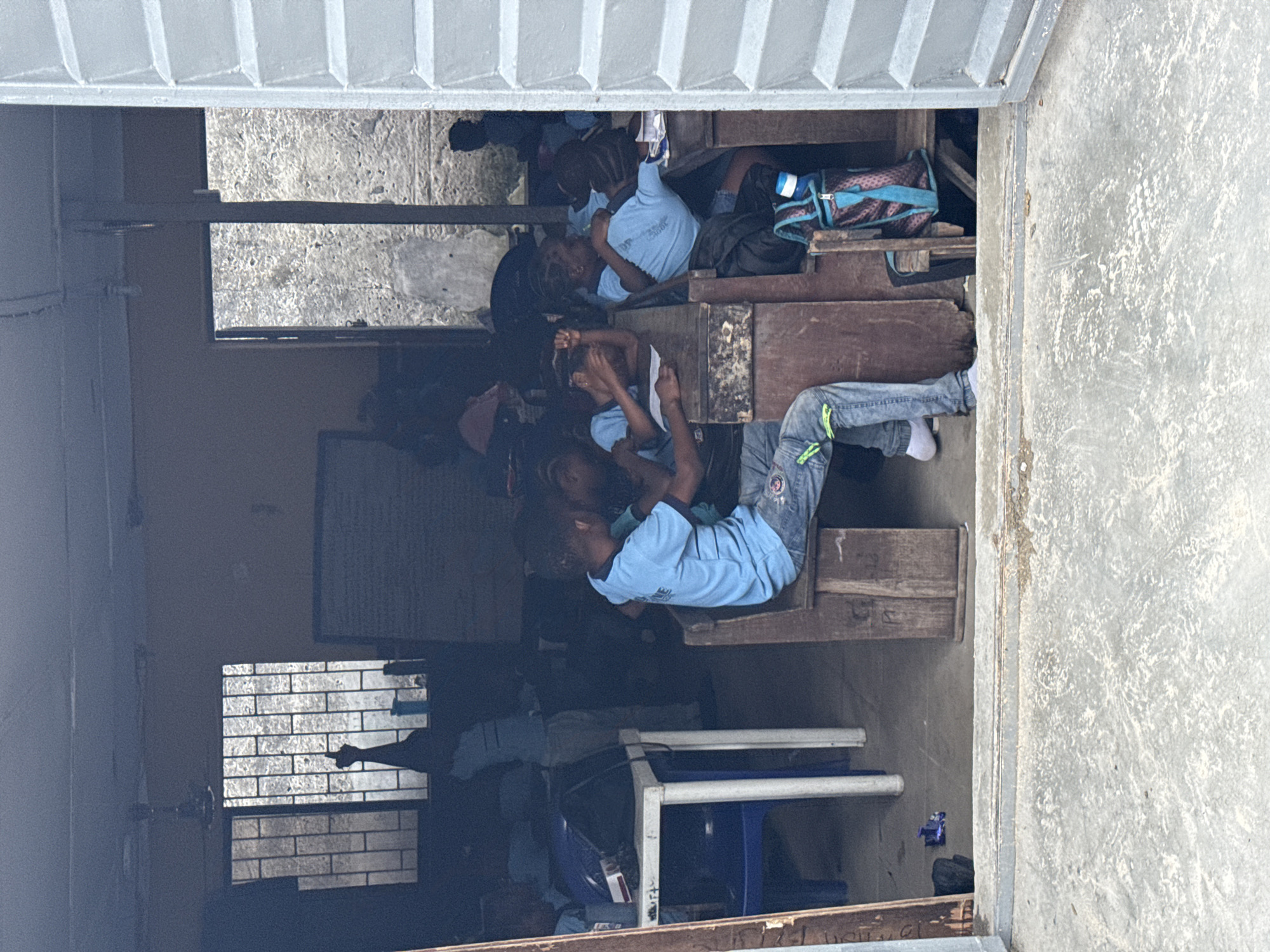 The image shows an outdoor classroom with several young students sitting on wooden benches. The students are wearing light blue shirts and some have backpacks. Some students appear to be resting or sleeping. The classroom has wooden desks and a whiteboard. There are also windows with metal bars. The scene appears to be set in a warm climate, possibly during the day, given the lighting. The overall atmosphere seems relaxed, with some children appearing to be taking a break. The image is captured from an angle looking into the classroom, with the top of the frame showing a corrugated metal overhang. The location context provided is Ikoyi, Nigeria. No visible text is discernible in the image.