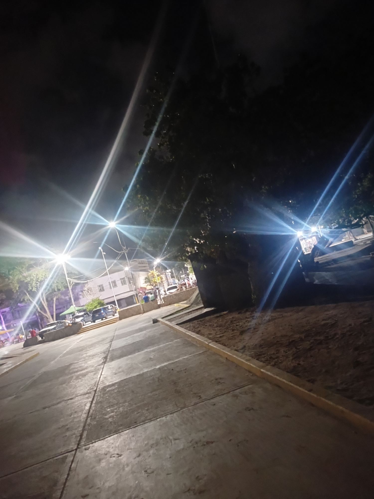 This is a nighttime photograph taken in Juan Griego, Venezuela, likely in a public square or street. The image is dominated by bright light flares from streetlights, creating strong streaks of light across the scene. The foreground shows a concrete sidewalk with distinct paving lines. To the left, there is a street or plaza area with several parked cars, including a white sedan and a dark blue minivan. A few people are visible in the background, some near the vehicles and others further in the distance, suggesting pedestrian activity. A building with multiple windows is visible behind the streetlights. To the right of the sidewalk, there is an area of dirt or sand, possibly a park or undeveloped land, bordered by a yellow curb. Trees with dark foliage are present in the mid-ground and background, partially obscuring the sky and buildings. The overall atmosphere is dimly lit, characteristic of a nighttime urban environment.