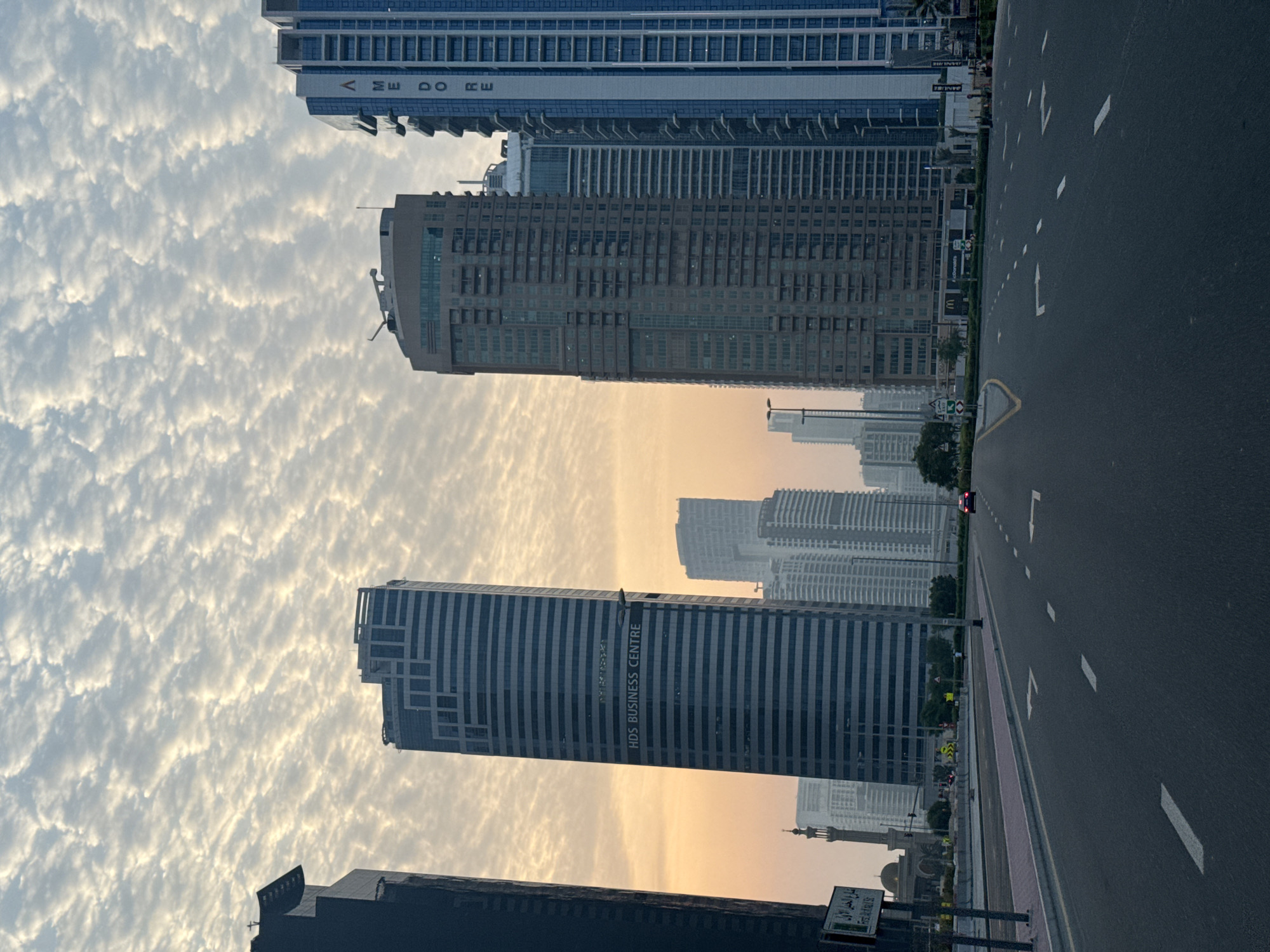 A street-level, eye-level, wide shot captures a cityscape at sunrise in Dubai, United Arab Emirates, with the sky filled with a pattern of altocumulus clouds. The foreground features an empty asphalt road with white lane markings and directional arrows. To the left of the road, a sidewalk and median separate it from a row of buildings and a mosque. A street sign is visible on a pole. Dominating the midground are several tall skyscrapers, including the HDS Business Centre with its name clearly displayed. Further back, more residential or commercial buildings form the urban landscape. The sun is low on the horizon, casting a warm glow on the sky and the upper portions of the buildings. A single red car is driving away from the camera on the road. There are no people in the immediate foreground or midground.