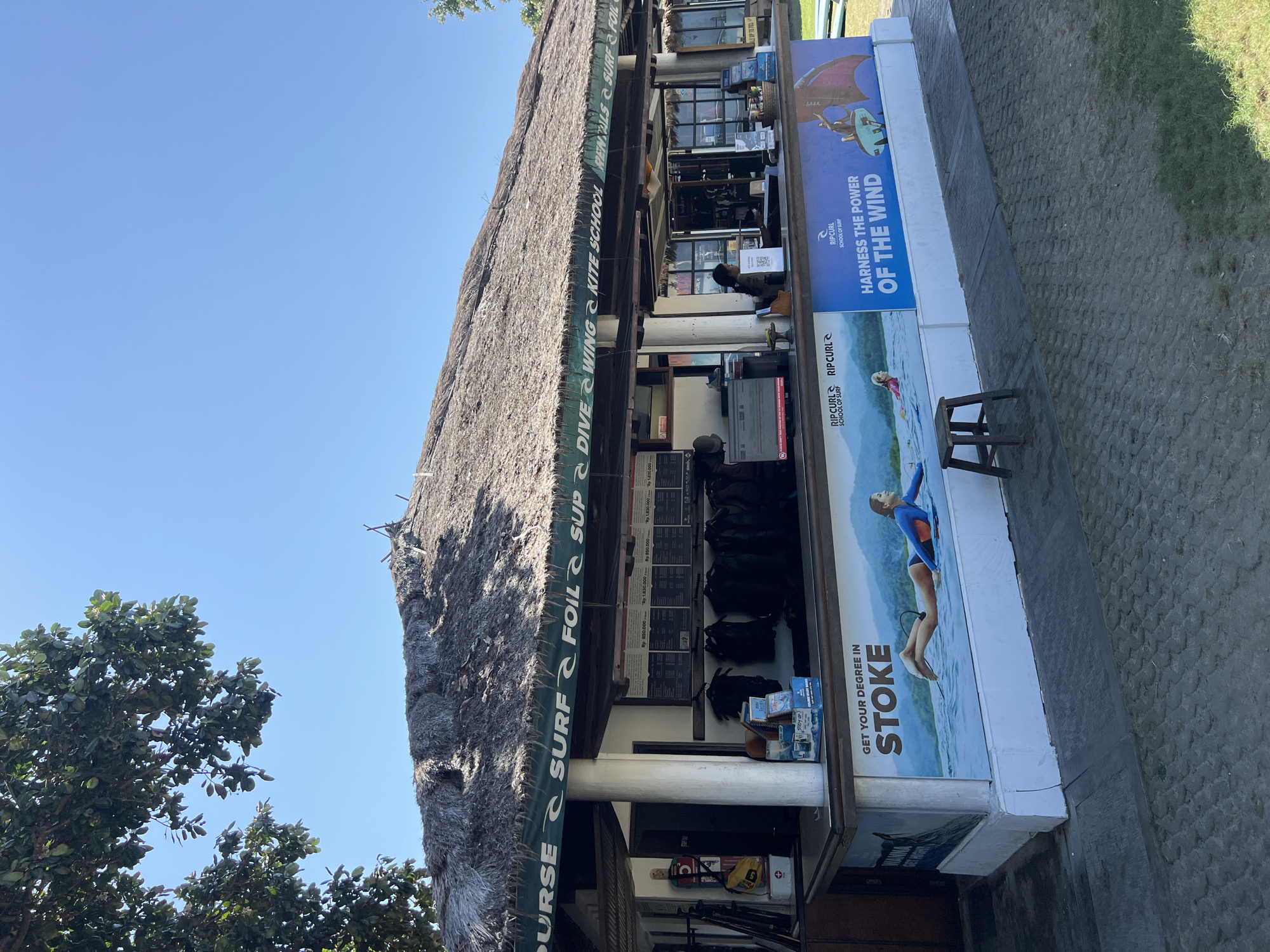 The image shows a Rip Curl surf school and rental shop with a thatched roof. A large banner wraps around the front of the building, featuring images of surfers and kitesurfers and promoting courses such as "Get Your Degree in Stoke" and "Harness the Power of the Wind." A price list for various courses and rentals is displayed inside the shop. Several wetsuits are hanging on display behind the counter, and surfboards and kitesurfing equipment are visible. A woman is working behind the counter, attending to customers. The shop appears to be located in Denpasar, Indonesia, given the Rip Curl branding and the tropical vegetation visible in the background. The ground in front of the shop is paved with cobblestones and sand.