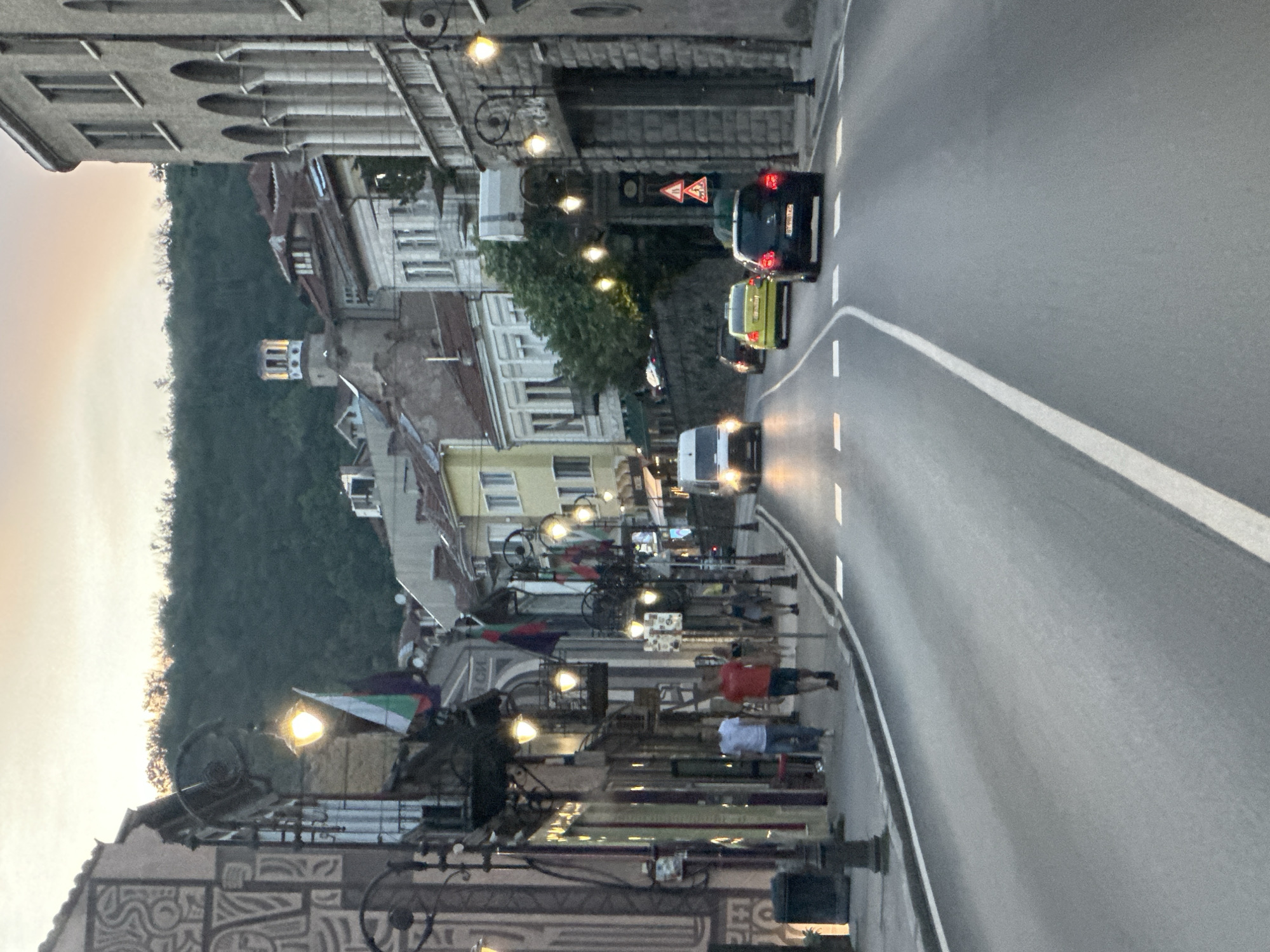 The image depicts a street scene in Veliko Tarnovo, Bulgaria, likely captured during dusk. The street, paved with asphalt, curves gently upwards into the distance, bordered by buildings on both sides. The architecture appears to be a mix of old-world European styles, with arched windows, balconies, and ornate facades. Several streetlights are illuminated, casting a warm glow on the scene.
In the foreground, a white van is approaching, its headlights on. Behind it, a yellow car and a black car are following. The road is marked with white lines, indicating lanes and pedestrian crossings.
On the left side of the street, pedestrians are walking along the sidewalk, some appearing to be tourists. Shops with illuminated windows are visible, suggesting commercial activity. Bulgarian flags are displayed on some buildings, hinting at the national context.
The background is dominated by a lush green hillside, densely covered with trees, rising towards a hazy sky. A distant tower, likely part of a historical building, is visible on the hilltop, illuminated by the setting sun. The overall atmosphere is serene and picturesque, with a sense of history and local charm.