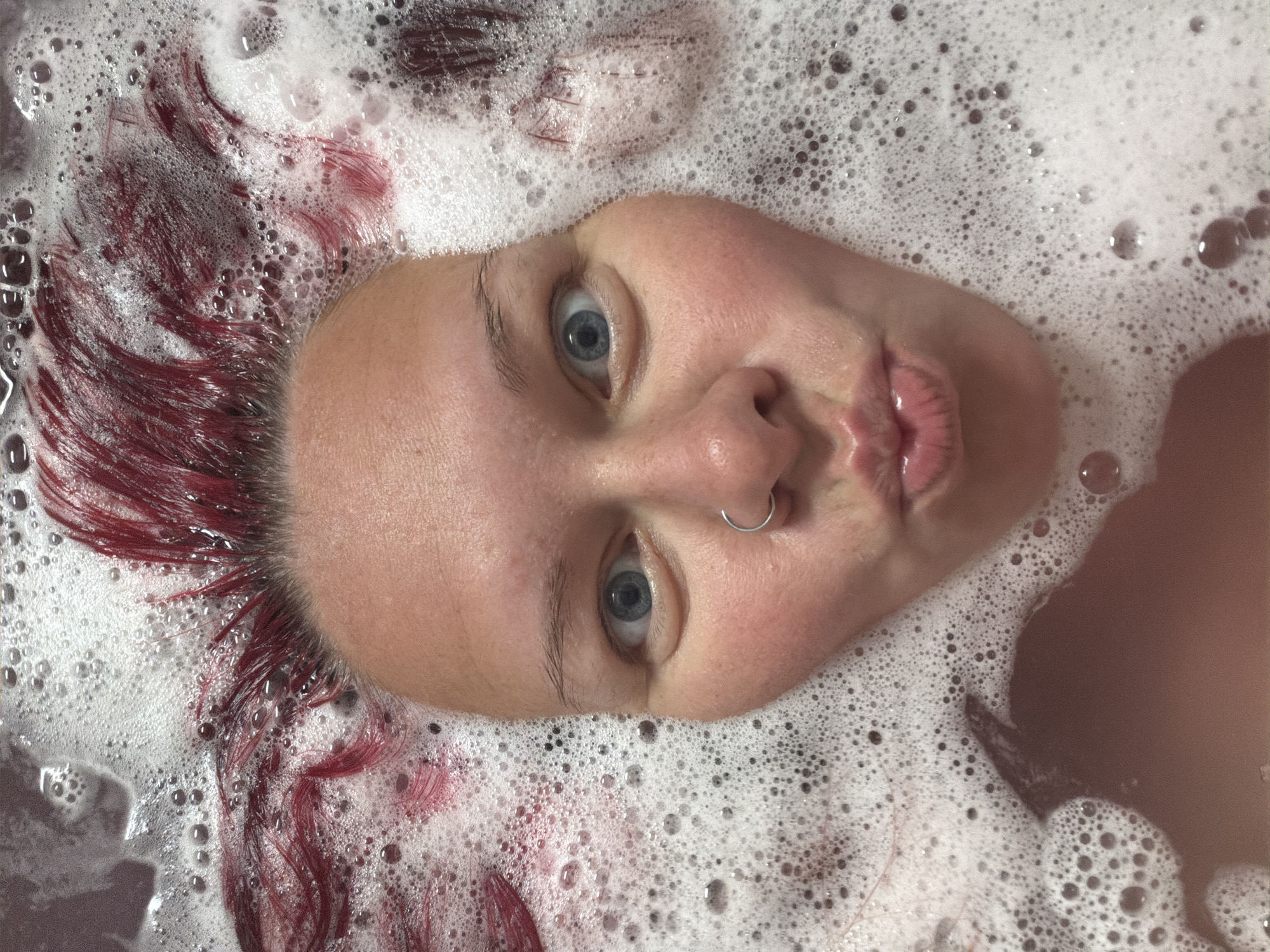 A close-up shot captures a person submerged in a bubble bath, with only their face and some of their red hair visible. The person's eyes are wide open, and their lips are pursed in a kissing gesture. A silver nose ring is visible on their right nostril. The bathwater is murky and reddish-brown, with a thick layer of white foam and bubbles covering most of the surface. The lighting is soft and diffused, creating a gentle glow on the person's skin. The scene appears to be indoors, likely in a bathroom. There is no information in the image to suggest its location in Chaikovsky, Russia.