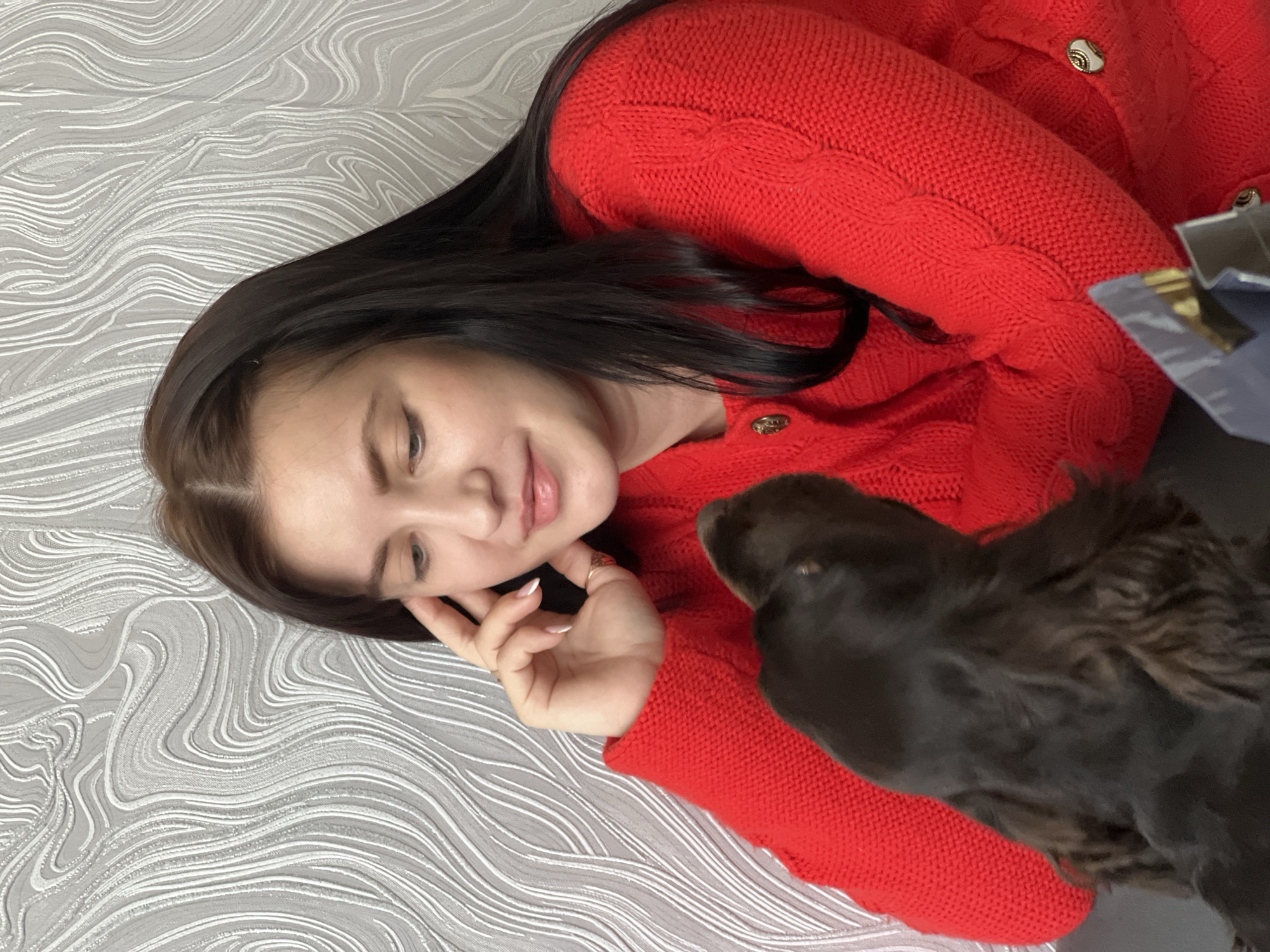 A woman with long dark hair, wearing a red cable-knit cardigan, is lying on a textured gray surface. Her head is turned towards the right, and she is looking down with a slight smile. Her right hand is resting near her face. A dark brown dog with long, floppy ears is close to her, its head partially obscuring her cardigan. The location is Марковский, Russia.