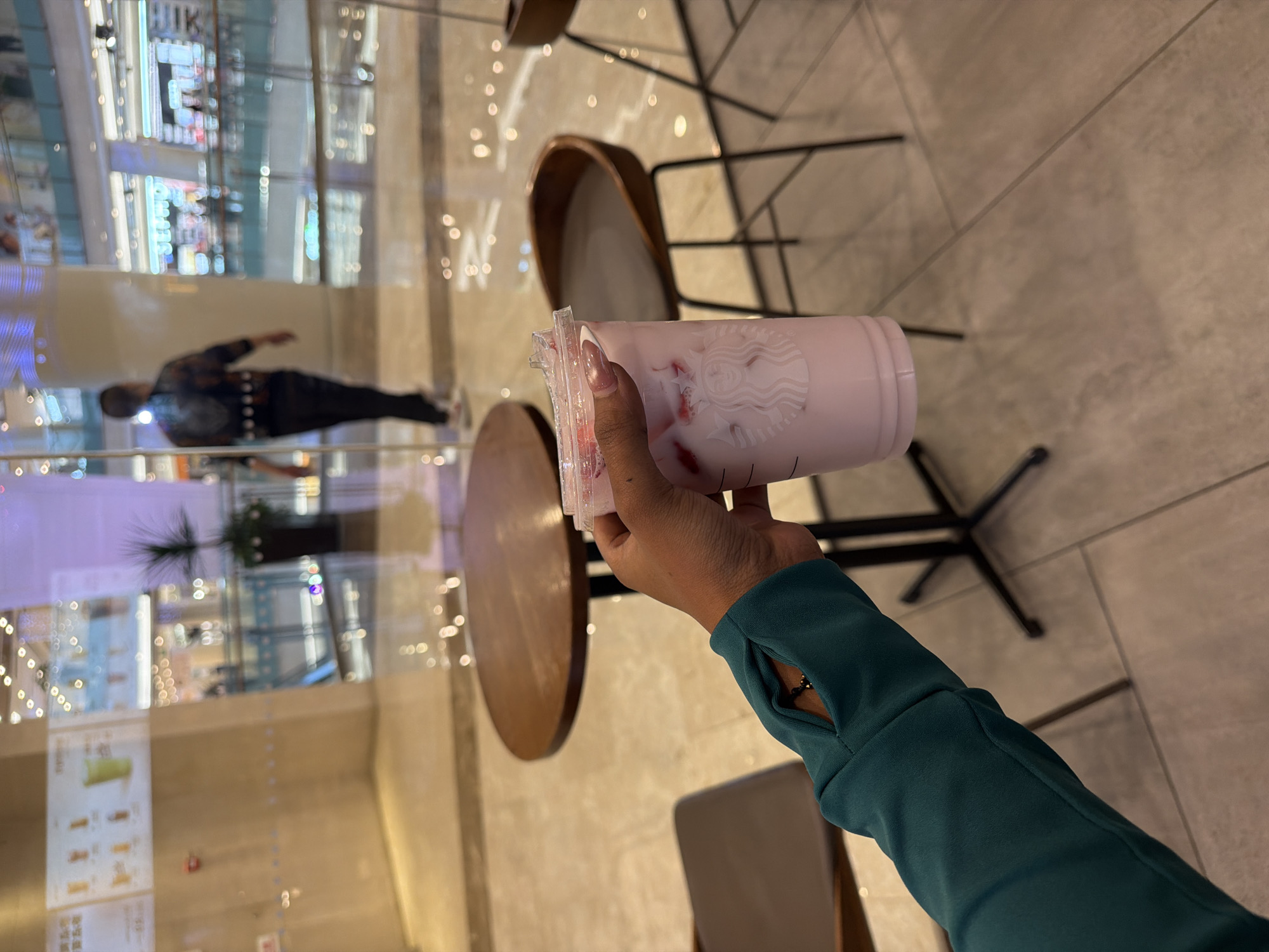 This image captures a first-person perspective shot within a modern indoor commercial space, likely a shopping mall or large complex in Yuhuatai District, China. The central focus is a hand holding a Starbucks cup filled with a light pink beverage containing visible fruit pieces, possibly strawberries. The person's hand has light-colored, possibly artificial, fingernails, and they are wearing a teal/dark green long-sleeved top with a thumbhole, revealing a black beaded bracelet or watch on their wrist. The Starbucks siren logo is clearly visible on the cup.
The background features a reflective glass wall or window, creating numerous bokeh light reflections and mirroring the interior environment. Through the reflections, one can discern blurry figures of people, including one person walking away from the camera, dressed in dark pants and a patterned top. Also visible in the reflections are parts of store displays, blurred architectural elements, and what appears to be a menu board with some unreadable text.
The setting includes brown wooden round tables and chairs with black metal frames, suggesting a cafe or seating area. The floor is a light-colored, possibly tiled surface with faint grid patterns. The space is brightly illuminated by artificial lighting, indicating an indoor environment where the time of day is not distinctly natural. The overall impression is that of someone taking a break to enjoy a drink in a bustling commercial hub.