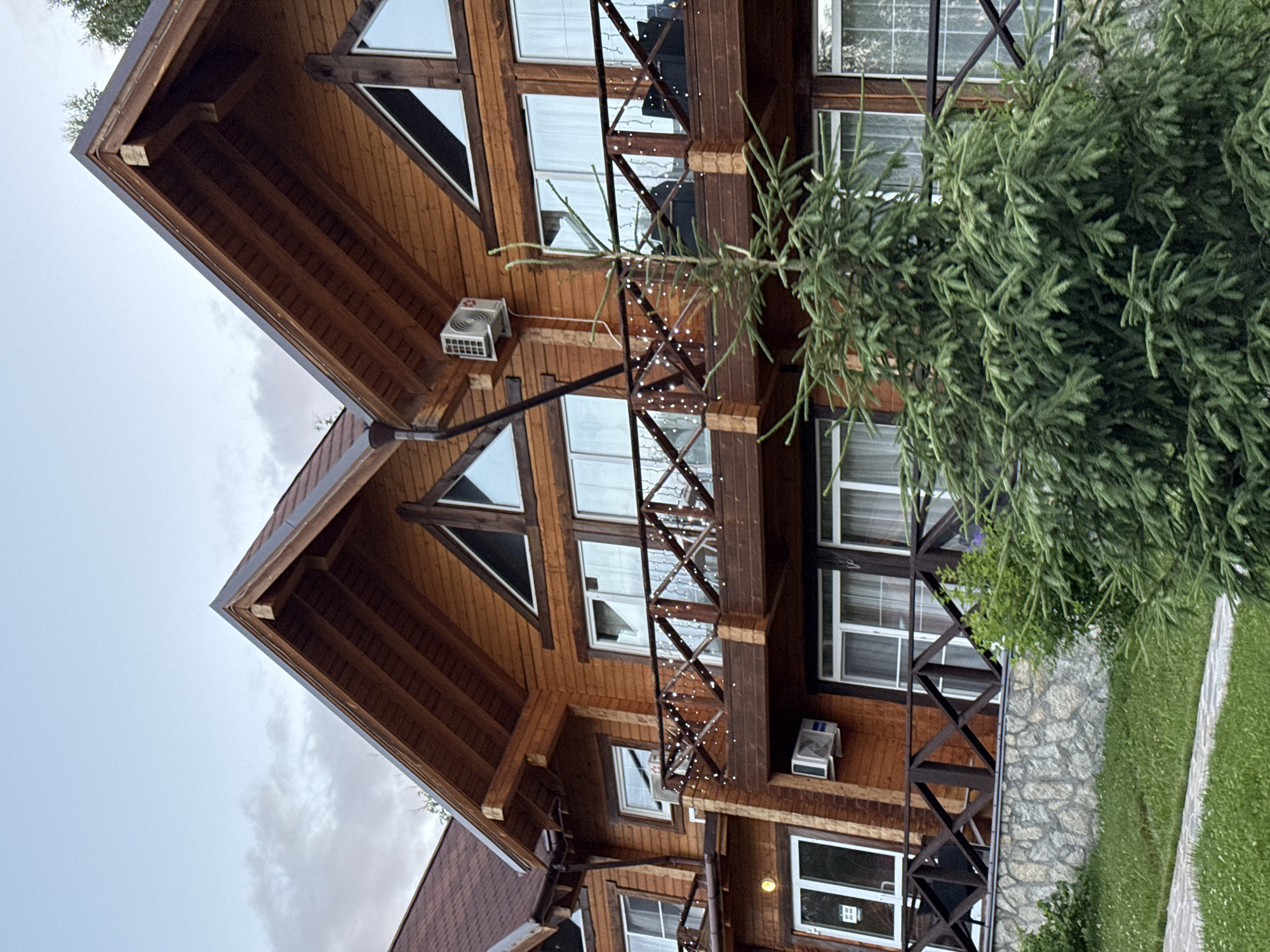 The image shows a multi-story wooden building with a dark brown roof and brown wooden siding. The building has several windows, including triangular windows on the upper levels and rectangular windows on the lower levels. A wooden balcony with a railing decorated with string lights runs across the second floor. Two outdoor air conditioning units are visible on the exterior walls. In the foreground, a large green evergreen tree is positioned in front of the building. The ground in front of the building is covered with green grass and a stone wall. The sky above is overcast. The location is identified as Syrostan, Russia.