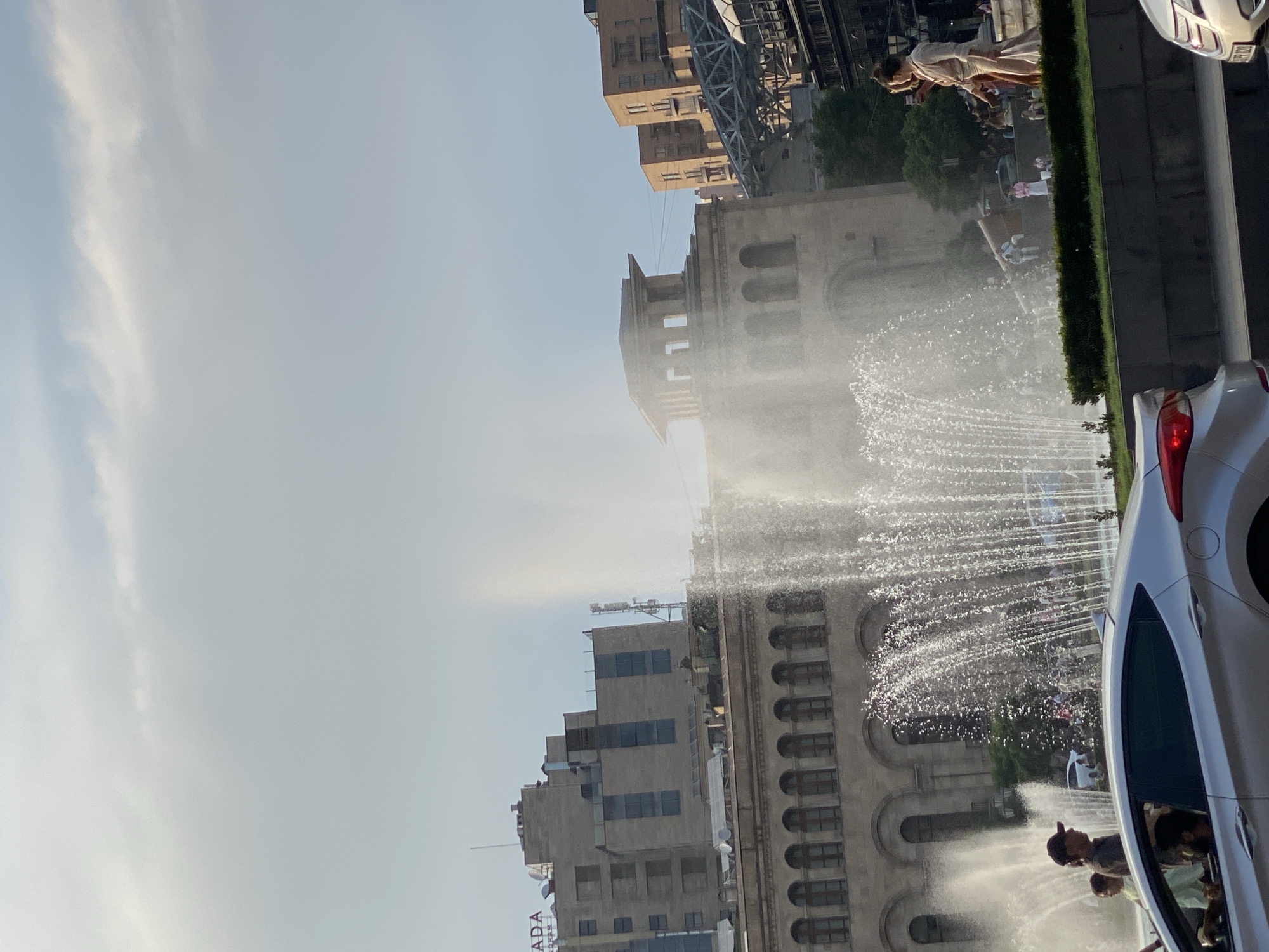 The image depicts a street scene in Yerevan, Armenia, likely at Republic Square, given the prominent architectural style of the buildings and the presence of a large fountain. The sky is overcast with scattered clouds. In the foreground, a silver sedan is parked or driving on a street, partially obscuring other elements. Behind the car, a fountain is actively spraying water, creating a misty atmosphere.
Several people are visible. On the left side of the fountain, two individuals, one wearing a dark baseball cap, are standing. Further back, more people are scattered around the fountain area and walking in the background. Two women are walking together on the right side of the fountain, one appearing to hold a phone.
The architecture in the background is characteristic of Soviet-era monumental buildings, with arched windows and stone facades. A multi-story building with the word "ADA" visible on its rooftop signage is to the left. To the right, other buildings with balconies and a modern metallic structure are visible. Lush green bushes border the fountain. The overall impression is an urban setting with public spaces, activity, and a grand architectural backdrop.