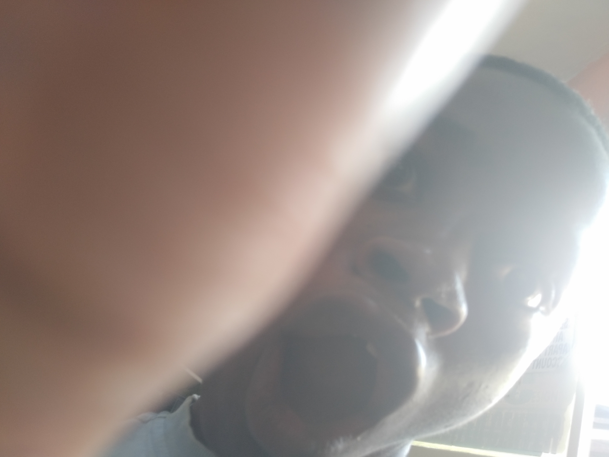 A close-up, poorly lit photograph shows a person with dark skin and an open mouth, likely a self-taken picture. The image is partially obscured by a blurry, large object in the foreground, possibly a finger or hand. The subject appears to be indoors, with ambient light and a blurred background suggesting an urban environment, specifically the city of Unknown, Unknown. Details of the scene are indistinct due to the proximity and poor lighting.