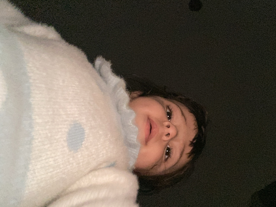 A person wearing a light blue and white sweater looks down at the camera from a low angle. The person's face, which includes brown eyes, a nose piercing, and brown hair, is centered. The background is dark and out of focus. The sweater has a fuzzy texture and a ruffled collar.