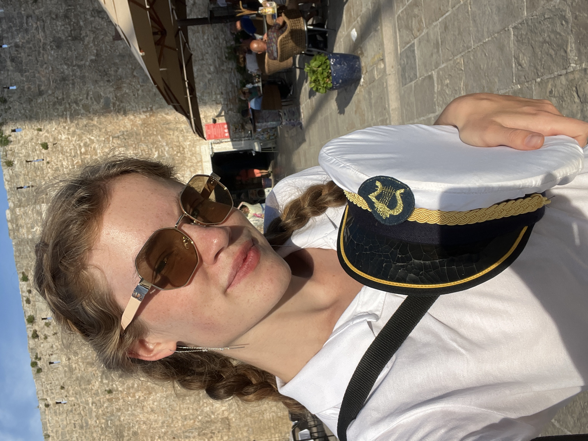 A fair-skinned woman with light brown hair in braids poses in what appears to be the city of Budva, Montenegro. She is wearing sunglasses and a white shirt with a black strap across her shoulder. She is holding a white sailor's cap with gold trim and a musical emblem on the front. In the background, there is a stone wall, a sun umbrella, and people sitting at tables, possibly at an outdoor cafe. A plant in a blue pot is also visible in the background.