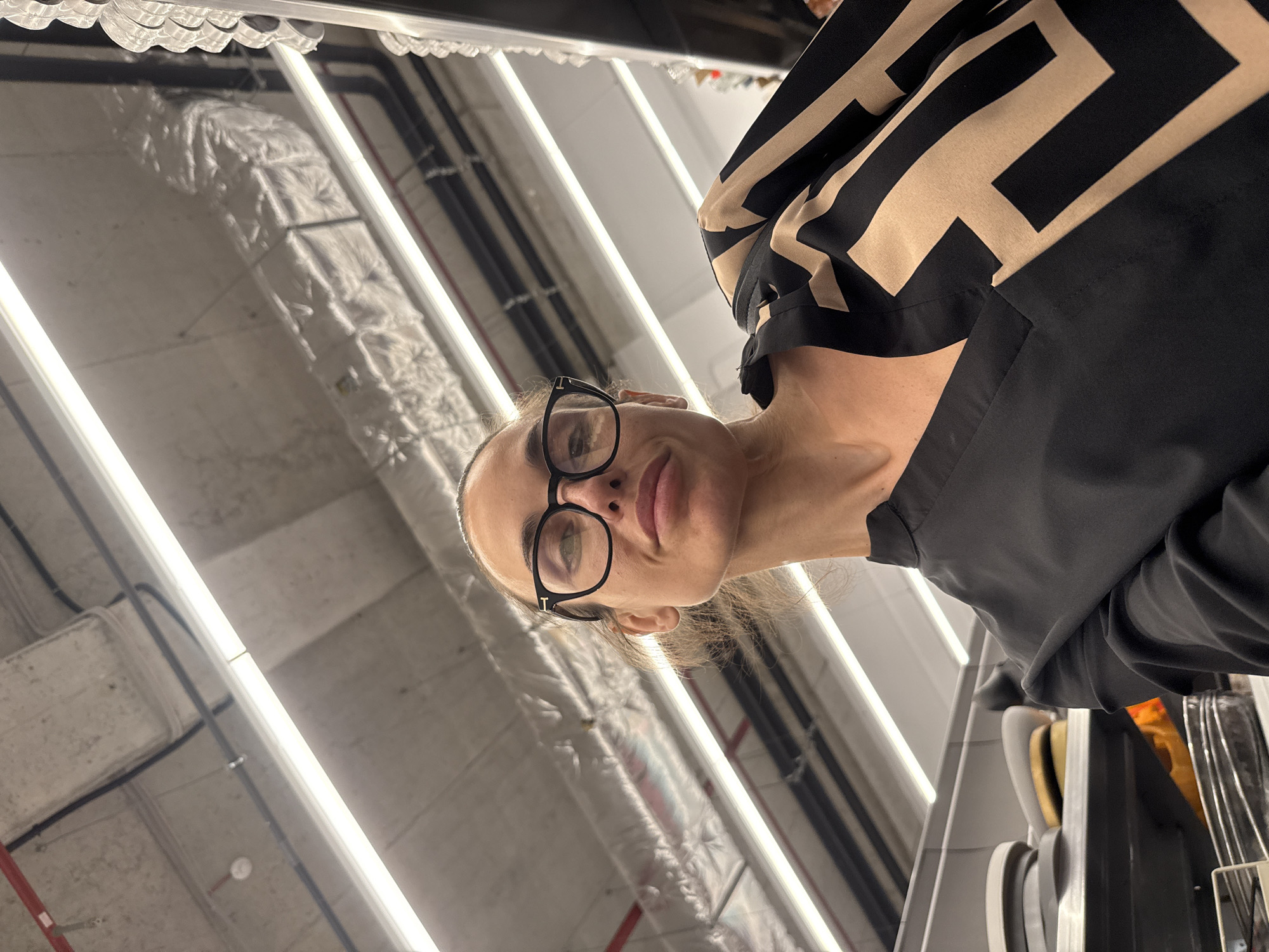 The image is a rotated indoor selfie, taken from a low angle looking upwards. The primary subject is a woman, captured from just above her nose to the top of her head, with a partial view of her right shoulder and arm. She has light skin, light-colored eyes (appearing green or hazel), and her hair is pulled back. She wears black-rimmed glasses with a distinct gold-colored 'T' logo visible on the right temple, likely indicating the brand Tom Ford. Her lips are slightly parted, and she has a neutral to slightly smiling expression. She is dressed in a top with a geometric black and beige pattern, and a plain black sleeve is visible on her right arm.
The background reveals a commercial or industrial interior ceiling. Bright, linear artificial lights run parallel across the ceiling, illuminating exposed utilities such as silver-insulated HVAC ducts, dark piping, and structural beams. A segment of a red pipe is also visible in one of the crops, possibly part of a fire suppression system. The ceiling itself appears to be made of concrete or a similar material. To the far right, obscured by the woman's shoulder and arm, there are hints of shelving or racks holding various items, suggesting a retail or warehouse environment. The overall lighting is bright and even, typical of such a setting. The time of day is not discernible due to the indoor artificial lighting. The location context provided is Pleven, Bulgaria.