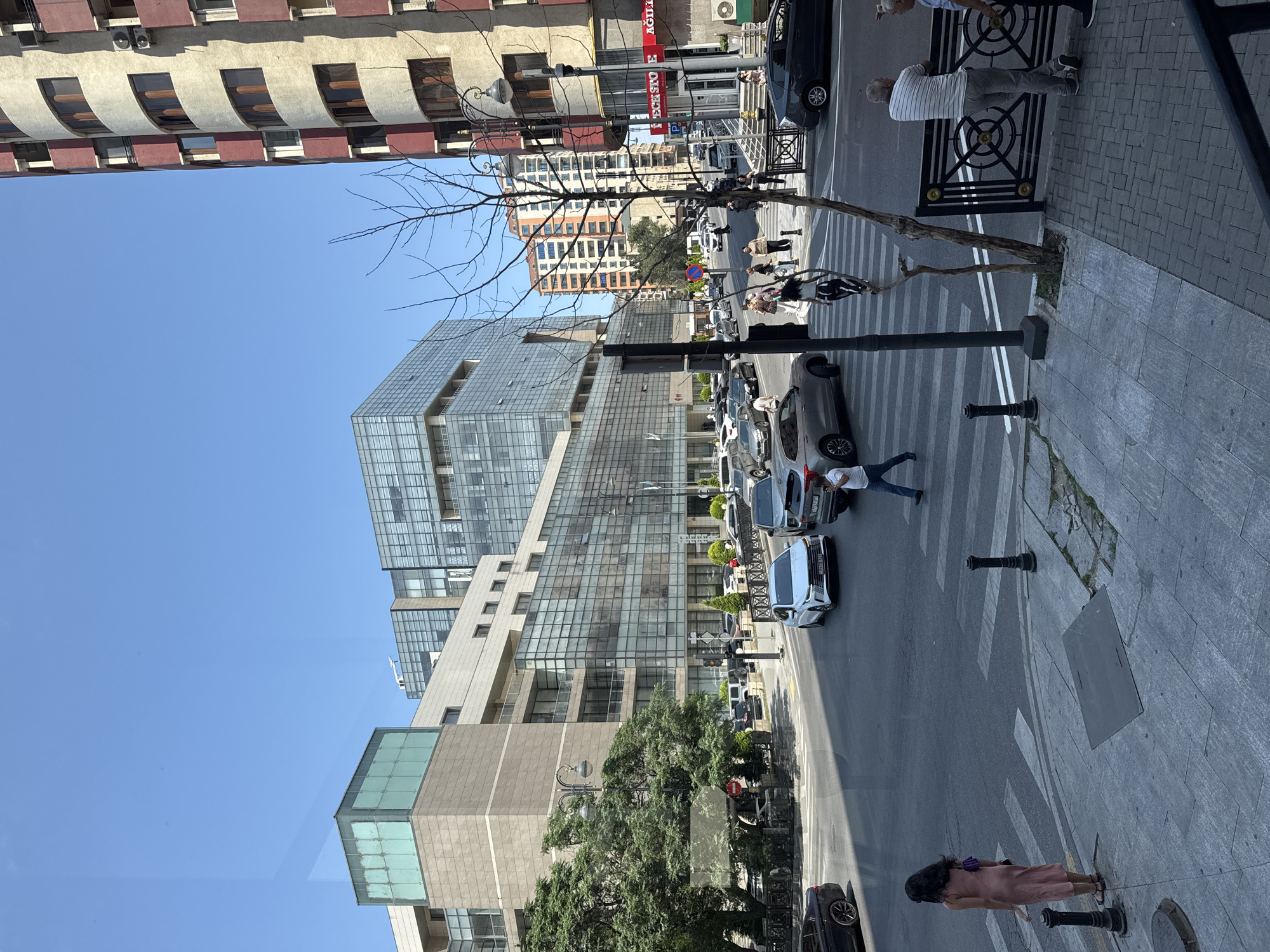 The image depicts a street scene in Baku, Azerbaijan, on a clear day. Multiple modern buildings with glass facades and balconies line the wide street. A multi-story residential building with a red and beige facade is visible on the right. Cars are driving on the road, and a pedestrian crossing is marked on the asphalt. Several individuals are present, including a woman in a pink dress standing on the sidewalk, a person in a white shirt and blue jeans walking across the pedestrian crossing, and a group of people near a fence on the right side of the image. Streetlights and trees are also part of the urban environment. The scene appears to be a busy intersection with a mix of commercial and residential structures.