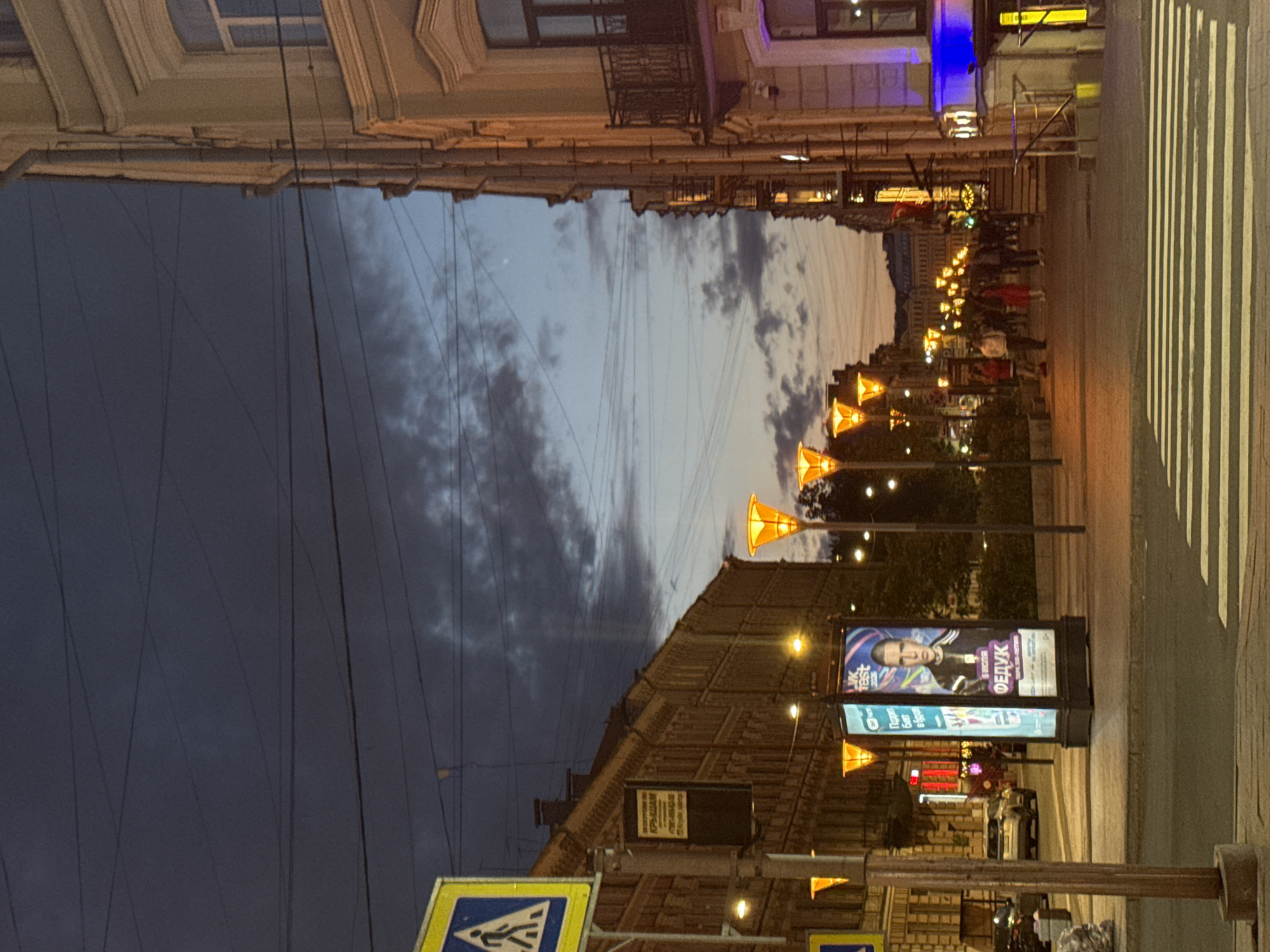 The image depicts a street scene in Saint Petersburg, Russia, at dusk. On the left side of the street, a multi-story building with intricate architectural details and numerous windows is visible. A pedestrian crossing is in the foreground, indicated by white stripes on the asphalt. A street sign featuring a pedestrian icon is mounted on a pole. Several ornate street lamps with bright orange light illuminate the scene. A large advertising billboard displays a portrait of a man and text in Russian.
On the right side of the street, another multi-story building with a light-colored facade, balconies, and ornate detailing is present. The sky is a deep blue, transitioning to lighter shades near the horizon, with dark clouds adding texture. Several people are walking along the sidewalk, some are conversing, and others are passing by. A car is parked on the left side of the street, and there are indications of businesses and shops with illuminated windows. Overhead, power lines crisscross the sky. The overall atmosphere is that of an urban environment at the end of the day.