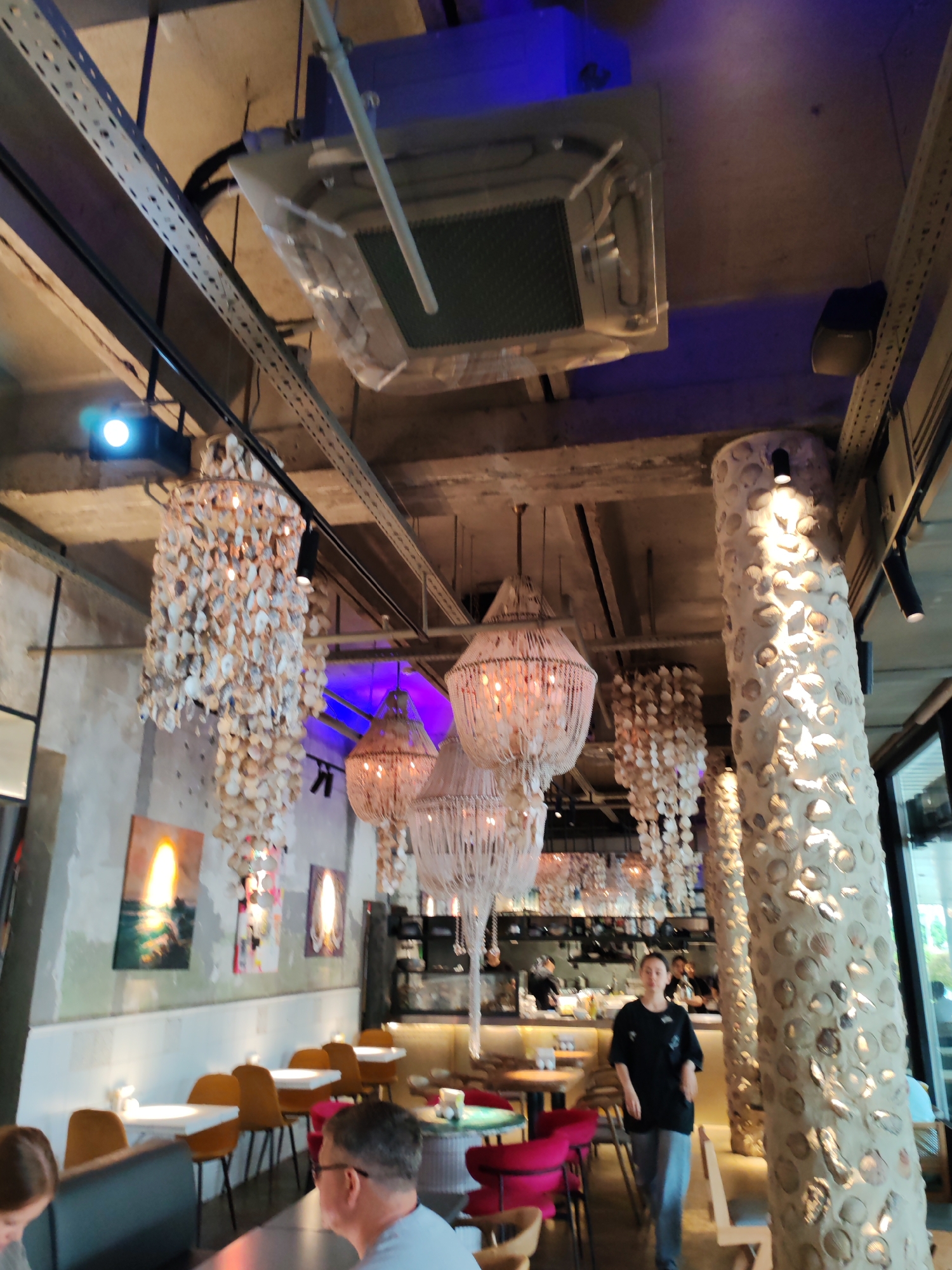 The image depicts the interior of a restaurant with a contemporary, bohemian aesthetic. The ceiling features exposed ductwork and lighting fixtures, including a large air conditioning unit covered in plastic sheeting. Several intricate chandeliers made of seashells hang from the ceiling, casting a warm glow. The walls are textured concrete, adorned with abstract paintings. The restaurant furniture includes white tables and mustard-yellow chairs, with some red chairs visible further back. A large, shell-encrusted pillar stands prominently in the foreground. A woman in a black t-shirt and grey pants is walking across the dining area, with other patrons and staff visible in the background near a bar and kitchen area. The overall ambiance suggests a trendy dining establishment, possibly located in Moscow, Russia, given the context provided.