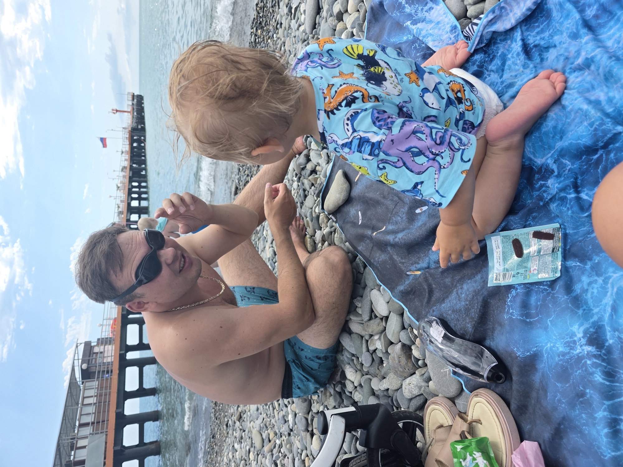 A man and a toddler are on a pebble beach in Sochi, Russia. The man, wearing sunglasses and a gold chain, is sitting on a blue towel and appears to be interacting with the toddler. He is wearing turquoise swim trunks with a pattern. The toddler, facing away from the camera, is wearing a light blue rash guard with an octopus print and a diaper. The toddler's feet are visible.
The scene appears to be a daytime outing, given the bright sky and sunlight. The weather seems pleasant, with a calm sea in the background and a pier with a Russian flag visible in the distance. The man's expression suggests a relaxed and happy mood, as he looks towards the toddler.
Objects visible include a blue towel with a wave pattern, a package of small dark brown snacks on the towel, a clear plastic item, a pair of light brown sandals, and a pink object. Part of a stroller is also visible in the foreground. The ground is covered in smooth, grey pebbles.