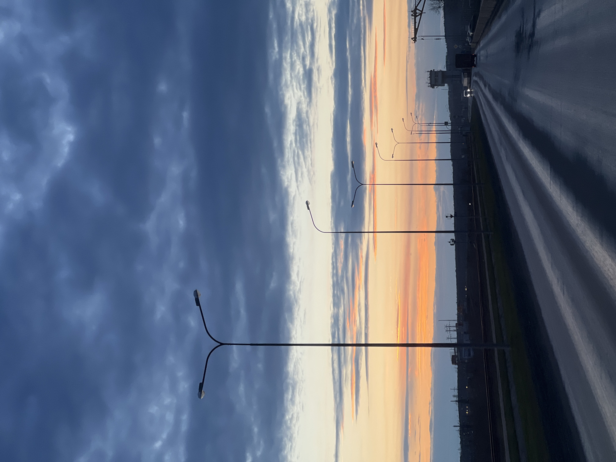 The image portrays a dramatic twilight scene along a wet road in Chaikovsky, Russia. The sky dominates the upper portion, showcasing a striking transition from dark blue-grey, somewhat ominous clouds on the left to vibrant streaks of orange and yellow light on the right, characteristic of either sunrise or sunset.
A wide, multi-lane asphalt road recedes diagonally into the distance on the right, its wet surface brilliantly reflecting the light in parallel, elongated streaks. In the far distance on the road, several vehicles are visible, including a dark truck and a car with illuminated headlights, navigating the damp conditions.
A series of modern streetlights with distinctive Y-shaped arms line the left side of the road, their tall, slender poles creating strong vertical elements against the expansive sky. Beyond the road to the right, a dark horizon reveals faint green vegetation and distant industrial or infrastructural structures, including utility poles and a building, with a prominent metal framework visible in the upper right corner.
The wetness of the road suggests recent rain or heavy dew, enhancing the reflective qualities of the scene. The atmosphere is one of serene transition, with the interplay of dynamic clouds and warm light. No people are visible, and there is no discernible text within the image.