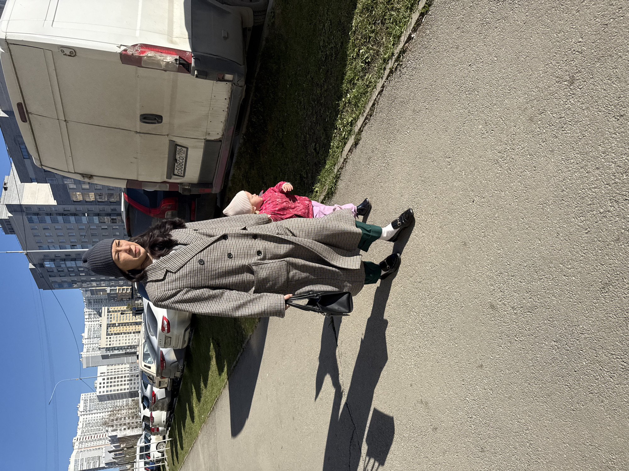 A woman in a gray houndstooth coat and a gray beanie stands on a sidewalk next to a white van. A child wearing a pink coat and a white hat lies on the woman's coat. The scene is set in Yekaterinburg, Russia, with high-rise buildings visible in the background. Cars are parked along the street. The sky is blue and sunny. The woman is holding a black purse.