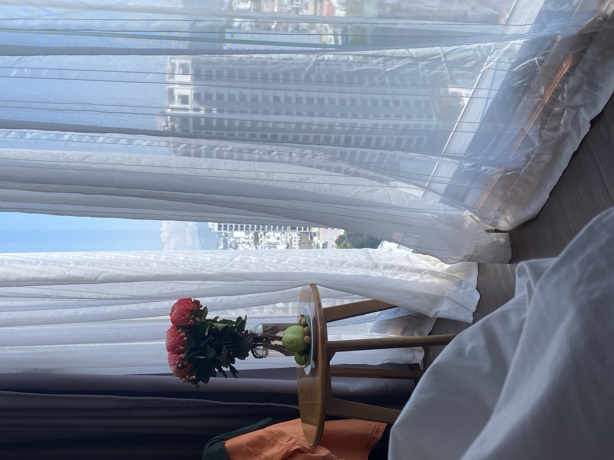 The image depicts a still life arrangement on a small wooden side table near a window in a room. A glass vase filled with a bouquet of red flowers stands on the table. Beside the vase is a small white plate holding two light green guavas and several small brown objects, possibly nuts or seeds. The table itself has three wooden legs and a round, light brown wooden top.
In the background, sheer white curtains are drawn, revealing a city view. The curtains are translucent, allowing the light to filter through and partially obscuring the buildings outside. Among the visible structures is a tall, modern building with a grid-like facade. The sky is a bright blue with some clouds.
To the left of the table, a dark gray curtain is visible, and to the left of that, an orange jacket is draped over something, possibly a chair or the edge of a bed. In the foreground, the corner of a white duvet or sheet is visible, indicating the presence of a bed. The floor appears to be wooden.
The scene is set in a room overlooking a cityscape, likely a hotel room or apartment in Nha Trang Ward, Vietnam, given the context. The overall mood is calm and domestic.