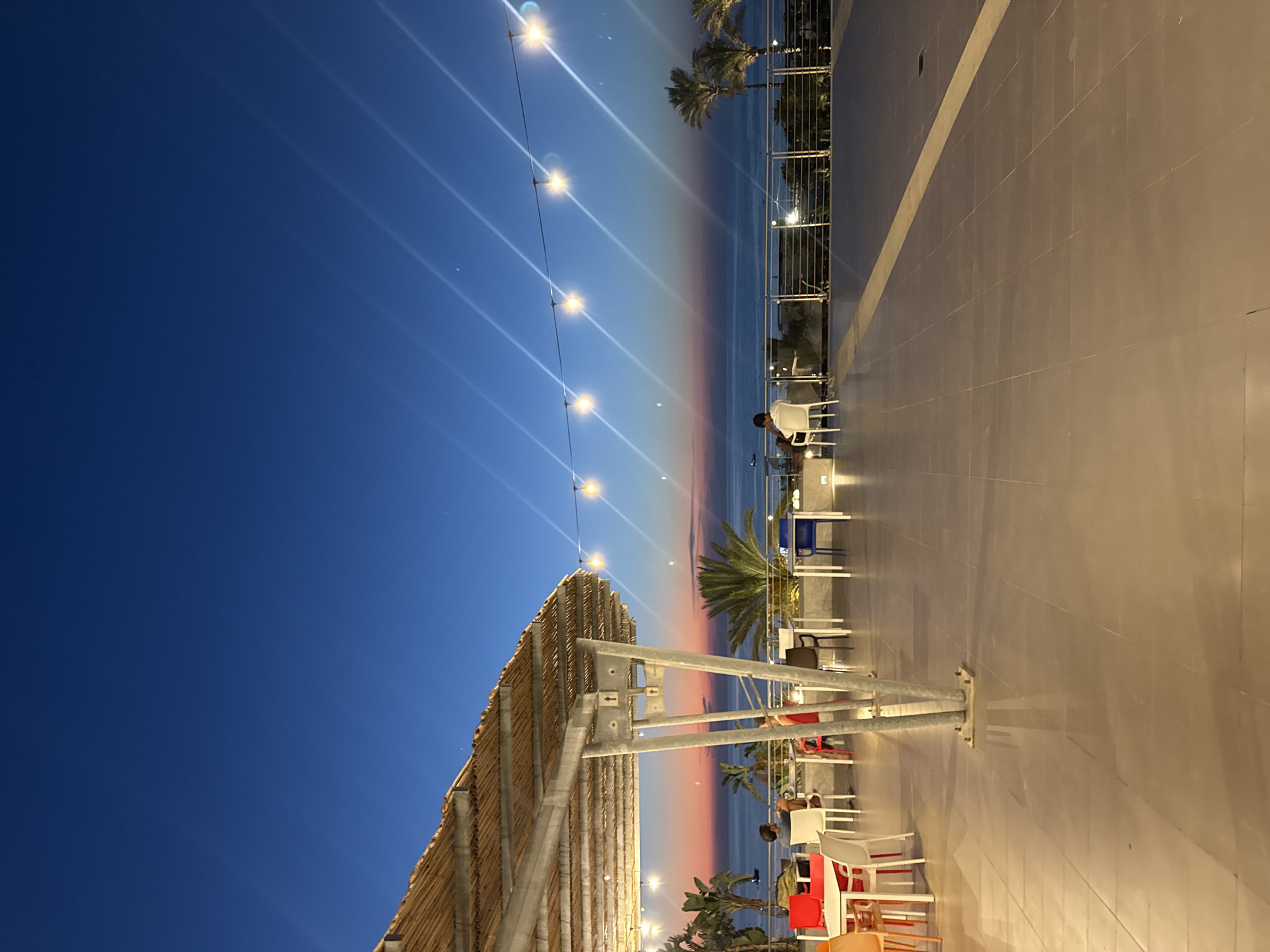 This image depicts a twilight scene at a restaurant overlooking the sea in Isola delle Femmine, Italy. The sky is a deep blue, transitioning to shades of pink and orange near the horizon where the sun has recently set. String lights with glowing bulbs are stretched across the upper portion of the frame, casting a warm light.
In the foreground, a tiled patio area features several tables and chairs, some occupied by individuals. To the left, under a straw-covered awning supported by metal poles, are white tables with red, white, and orange chairs. Further back, more tables and chairs are visible, with some patrons seated. A man in a white shirt and dark shorts is seated at a table on the right, facing the sea. Another person in a red top is seated at a table in the middle ground.
The middle ground shows a railing overlooking the sea, with palm trees visible to the right. The sea itself is calm and reflects the colors of the sunset. The background includes a faint outline of buildings and more palm trees. The overall atmosphere is one of a relaxed evening by the coast.