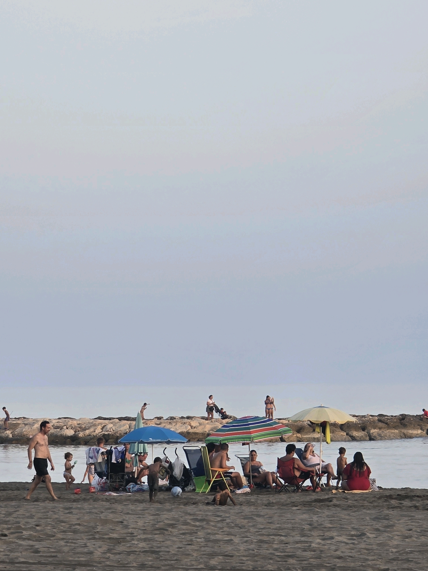 The image depicts a beach scene with several people enjoying leisure time on a sandy shore. In the foreground, a man walks to the left, while children play on the sand. Further back, groups of people are seated under beach umbrellas, some in chairs and others on towels. A rock barrier extends into the water, with individuals visible on top of it. The sky is overcast and the sea is calm. This scene is likely from a beach in Málaga, Spain, known for its coastal attractions.