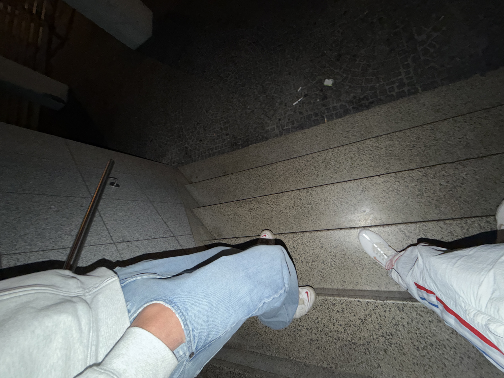 A high-angle, artificial-light photograph captures two individuals on a speckled light grey stone staircase at night in Vila Real, Portugal. The upper individual, visible from the mid-torso, wears a light grey sweatshirt, light blue loose-fit denim jeans with a tear near the right thigh pocket where a hand is placed, and white athletic shoes with red accents. The lower individual, visible from the lower torso, wears white track pants with vertical red and blue stripes along the sides and white athletic shoes with orange/tan and red accents. A metallic handrail is affixed to the left wall, which features light grey, textured panels. The bottom right shows a section of dark, traditional Portuguese cobblestone pavement, on which several white cigarette filters and a small white rectangular object are scattered. The background is dark and indistinct, with faint structural outlines.