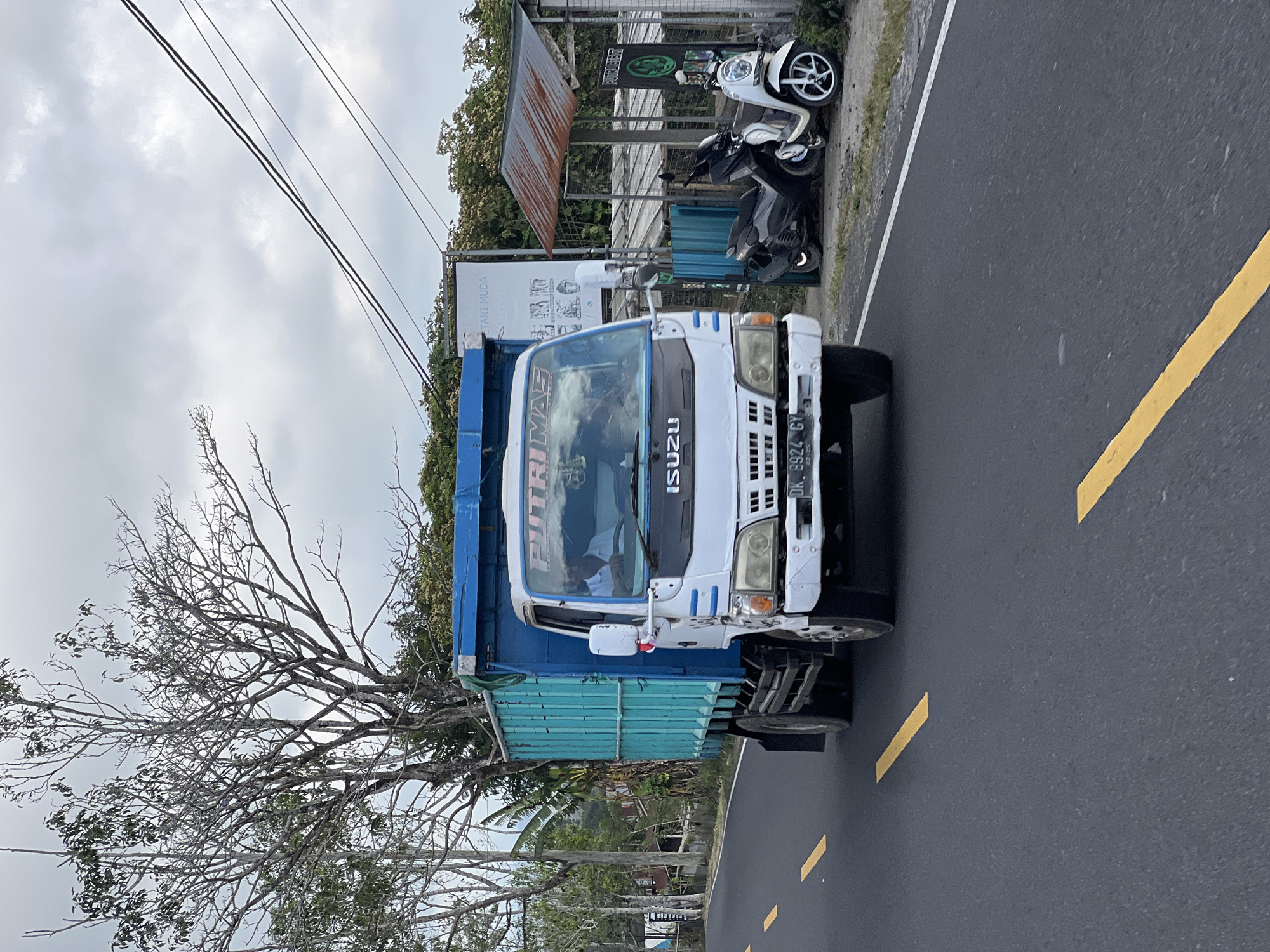 A white Isuzu truck with a blue cargo box drives on a paved road in Kintamani, Indonesia. The truck has a partial view of its driver, a man in a white shirt, behind the windshield. The truck's license plate reads "DK. 8924 GY". To the right of the truck, two motorbikes are parked next to a small shelter with a corrugated metal roof. In the background, trees and utility poles line the road. The sky is overcast.