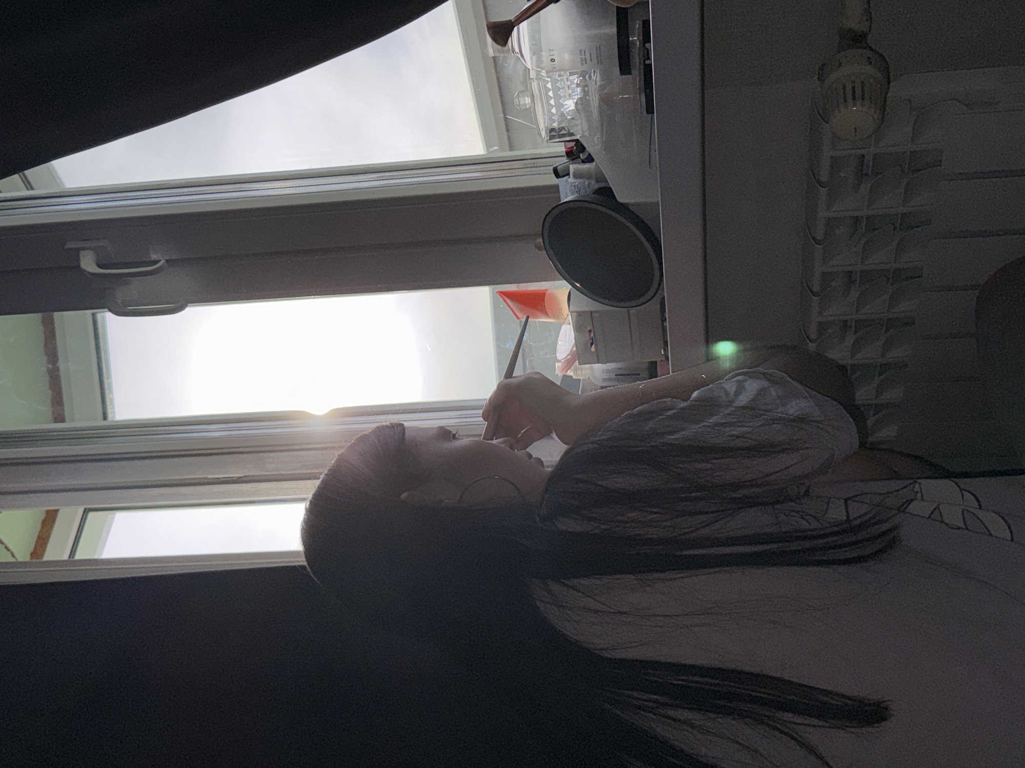 A woman lies horizontally on a bed in a room in Chaikovsky, Russia. She is positioned against a window with sunlight shining through. She appears to be applying makeup with a brush. The room contains a radiator and cosmetic items on a surface near the window. The lighting in the room is dim, except for the bright sunlight coming through the window. The photo is oriented at an angle.