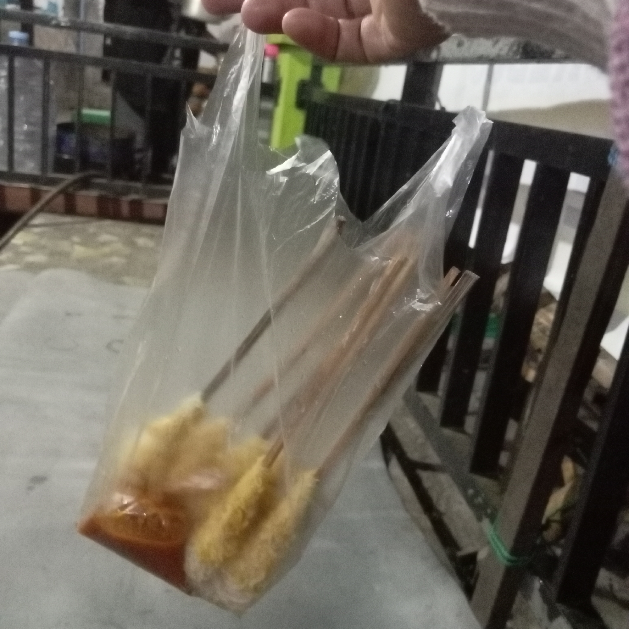 A person's hand, with visible fingernails and knuckles, holds a clear plastic bag filled with what appears to be food items on skewers. The food is coated in a yellow, textured substance and partially submerged in a reddish sauce at the bottom of the bag. The bag is slightly crumpled and shows signs of moisture. In the background, a dark-colored, slatted fence or railing is visible, along with some blurred structures and a glimpse of a light-colored surface. The scene suggests an outdoor or semi-outdoor setting, possibly a street food vendor or a casual dining environment. The image is taken from a slightly high angle, looking down into the bag.