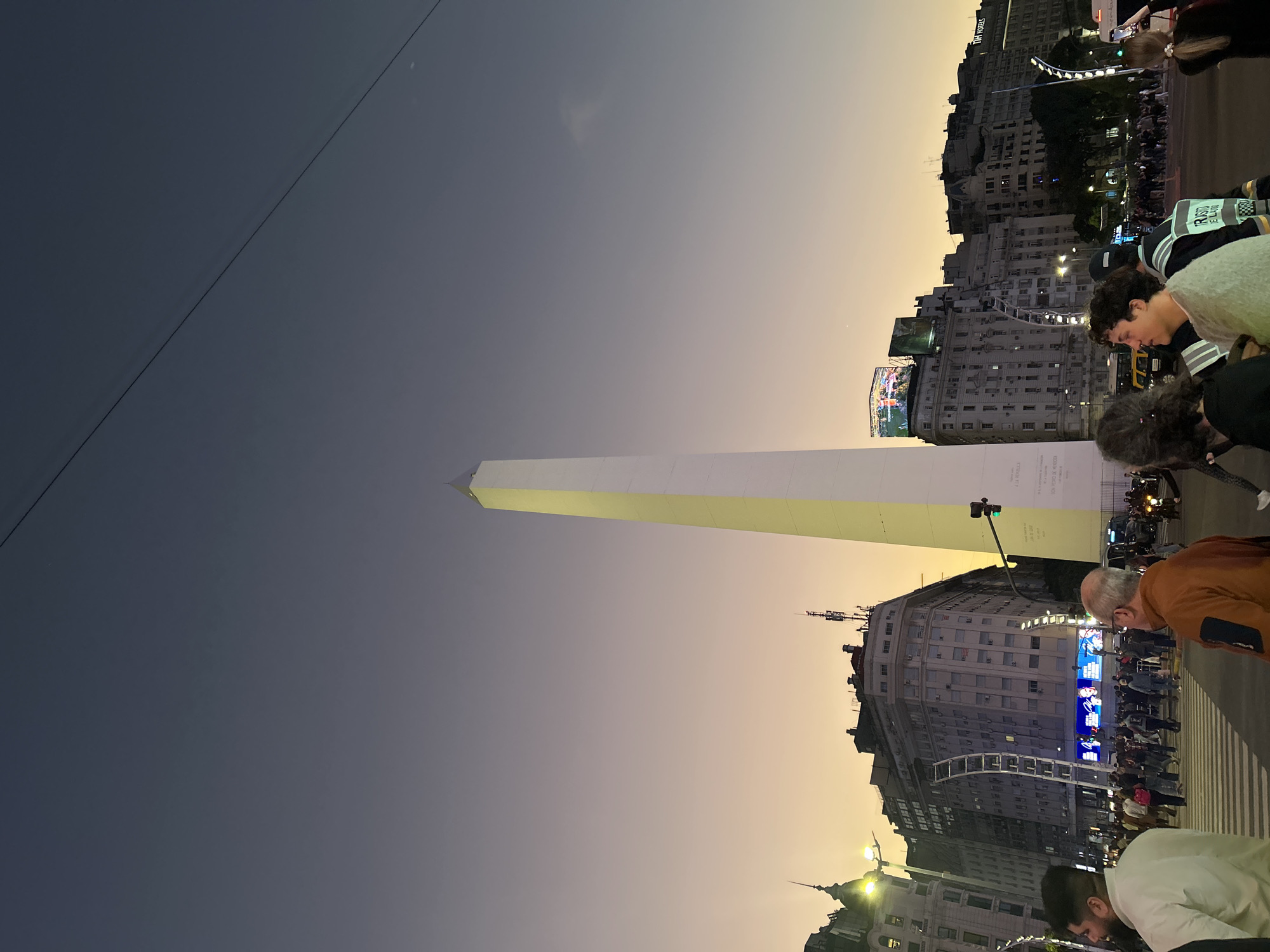 This is an eye-level, outdoor, nighttime shot taken in the city of Buenos Aires, Argentina, featuring the Obelisco monument. The obelisk appears to be a tall, slender, white structure. The sky in the background is a gradient of hues, ranging from dark blue to orange, indicative of either dusk or dawn.
Several multi-story buildings flank the obelisk on either side. Some buildings have lit signs and windows, suggesting activity. People are standing and walking in the foreground, with some engaged in conversation. There are also utility poles and traffic lights visible. The scene indicates an urban environment with pedestrians.