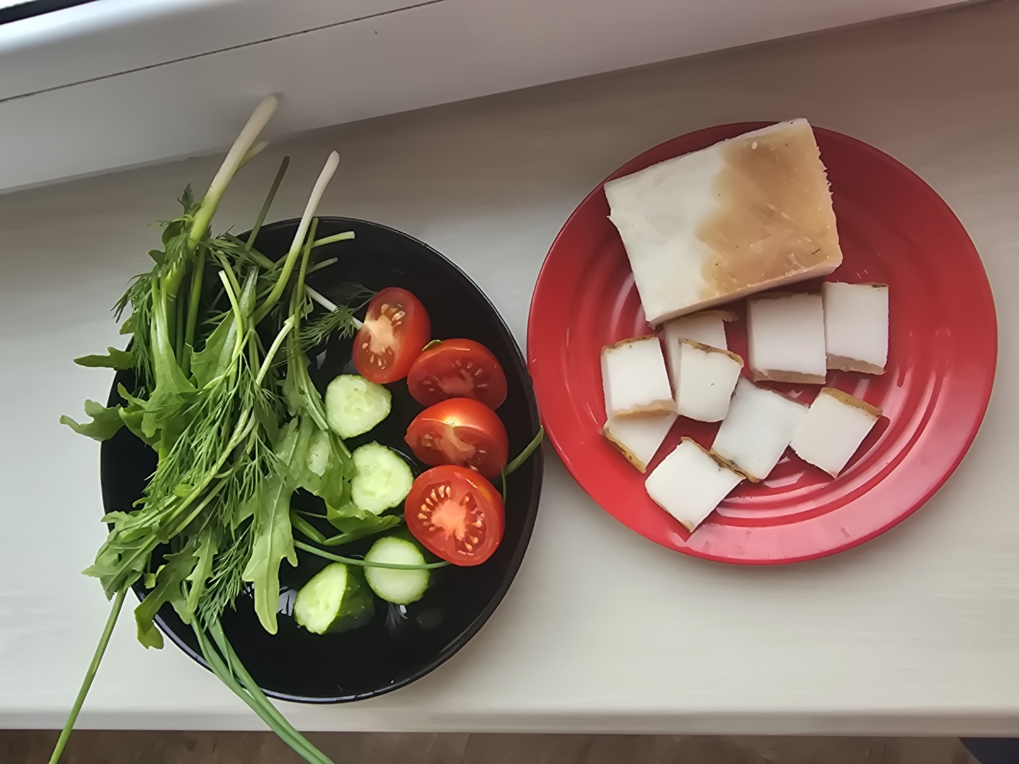 An overhead, high-angle shot shows two plates of food on a light-colored, possibly wooden surface. The top plate, black and round, contains a salad composed of arugula, dill, sliced green onions, sliced cucumbers, and halved cherry tomatoes. The bottom plate, red and round, holds several pieces of cut bacon and one larger piece of bacon. The lighting suggests an indoor setting, possibly a kitchen or dining area. The scene is devoid of any human presence. Based on the provided information, this image is from Sofiivska Borshchahivka, Ukraine.