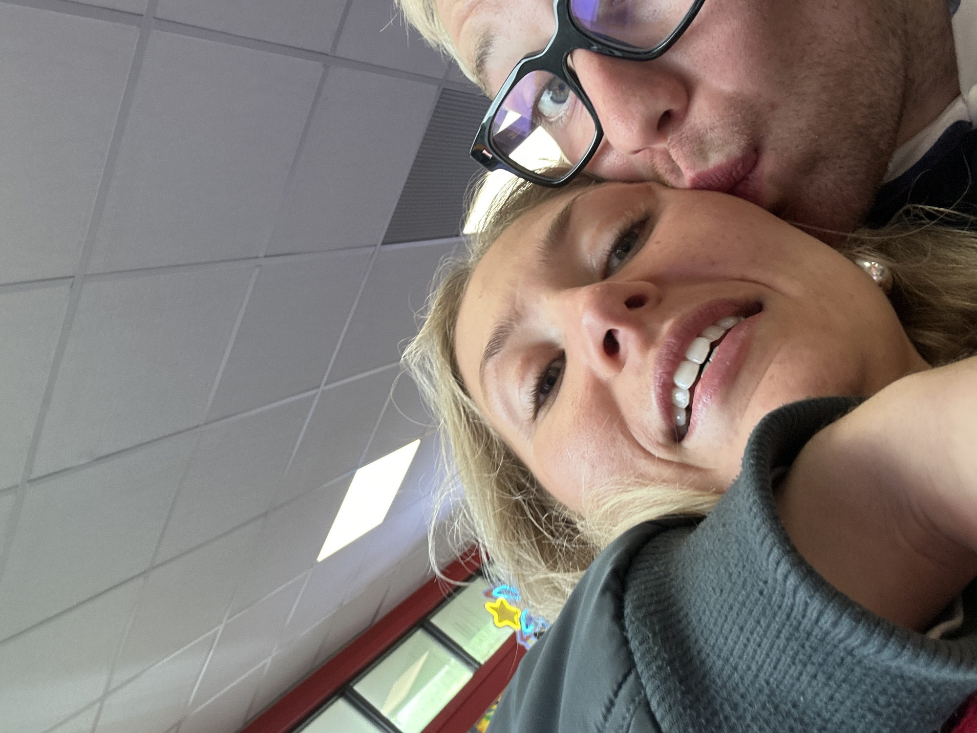 A close-up, upward-angled image depicts two individuals, likely a couple, engaged in an affectionate interaction within an indoor setting in Great Yarmouth, United Kingdom. On the left, a woman with blonde hair and light skin smiles directly at the camera, her teeth visible. She wears a dark jacket, with a ribbed cuff visible on her left arm, and an earring. On the right, a man with light hair and stubble, wearing black-framed glasses, kisses her left cheek. His eye is visible behind the glasses, also directed towards the camera. The background features a suspended grid ceiling with white panels and embedded, illuminated square or diamond-shaped light fixtures. To the upper left, a red structural element is visible alongside windows with dark frames. A neon sign shaped like a yellow star with a blue outline is partly visible behind the woman's head.