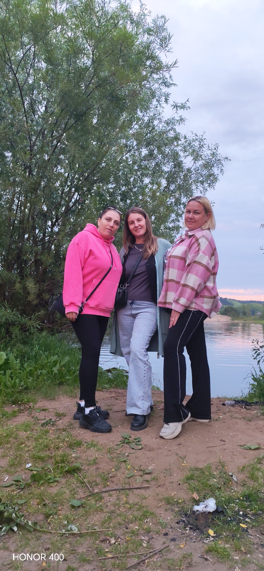 Three women stand in front of a large tree and a body of water. The woman on the left wears a pink hoodie and black leggings. The woman in the middle wears a gray t-shirt, a light blue cardigan, and light-wash bell-bottom jeans. The woman on the right wears a pink and white plaid shirt and black pants. The background shows a lake or river, trees, and a cloudy sky. The ground is covered with dirt and grass. The location is identified as Noviy, Russia.