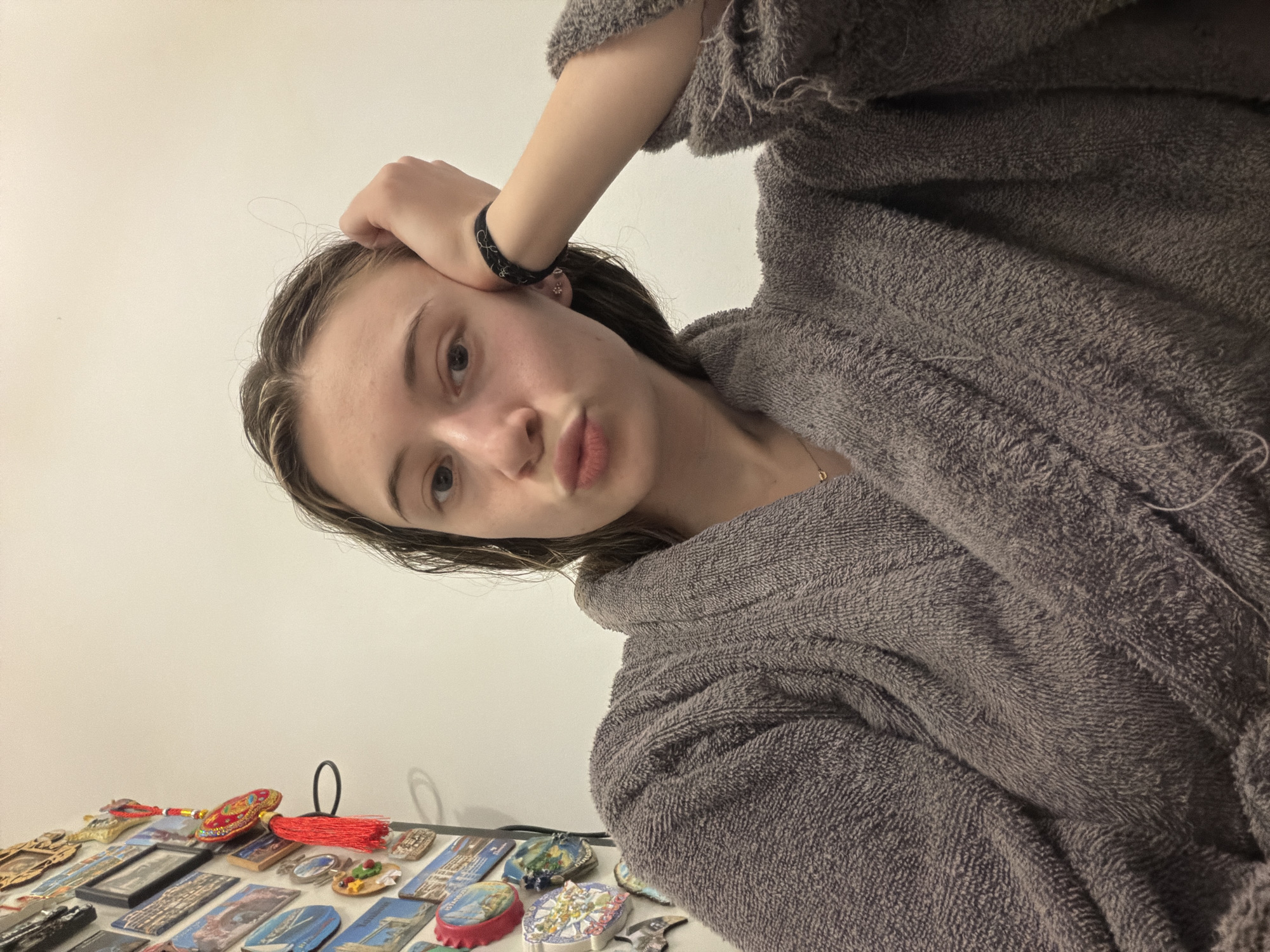 A person wearing a grey, textured robe is positioned in the upper portion of the frame. The individual has their hand raised to their head, and is looking directly at the camera with pursed lips. A dark bracelet is visible on their wrist. Below, a white surface holds a collection of souvenirs, predominantly magnets. The subjects of the magnets vary, with some depicting cityscapes, while others have decorative or artistic designs. The background appears to be a plain, off-white wall. There is no indication of the location being in Plovdiv, Bulgaria.