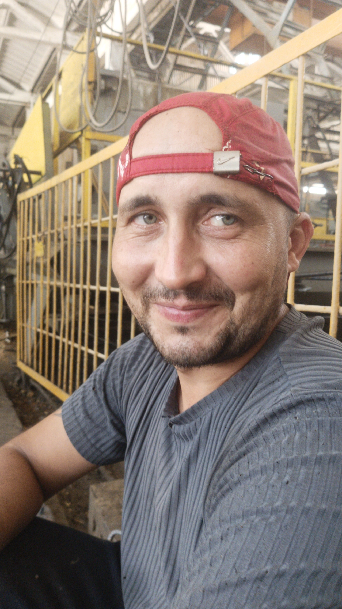 A man with a red baseball cap and a grey t-shirt is shown in a close-up portrait. The man has light green eyes, a mustache, and a beard. He is smiling and looking at the camera. The background appears to be an industrial setting, possibly a factory or workshop, with yellow metal bars and machinery. The image is taken indoors, with overhead lighting. There is no specific indication of the city of Gulkevichi, Russia within the image itself.