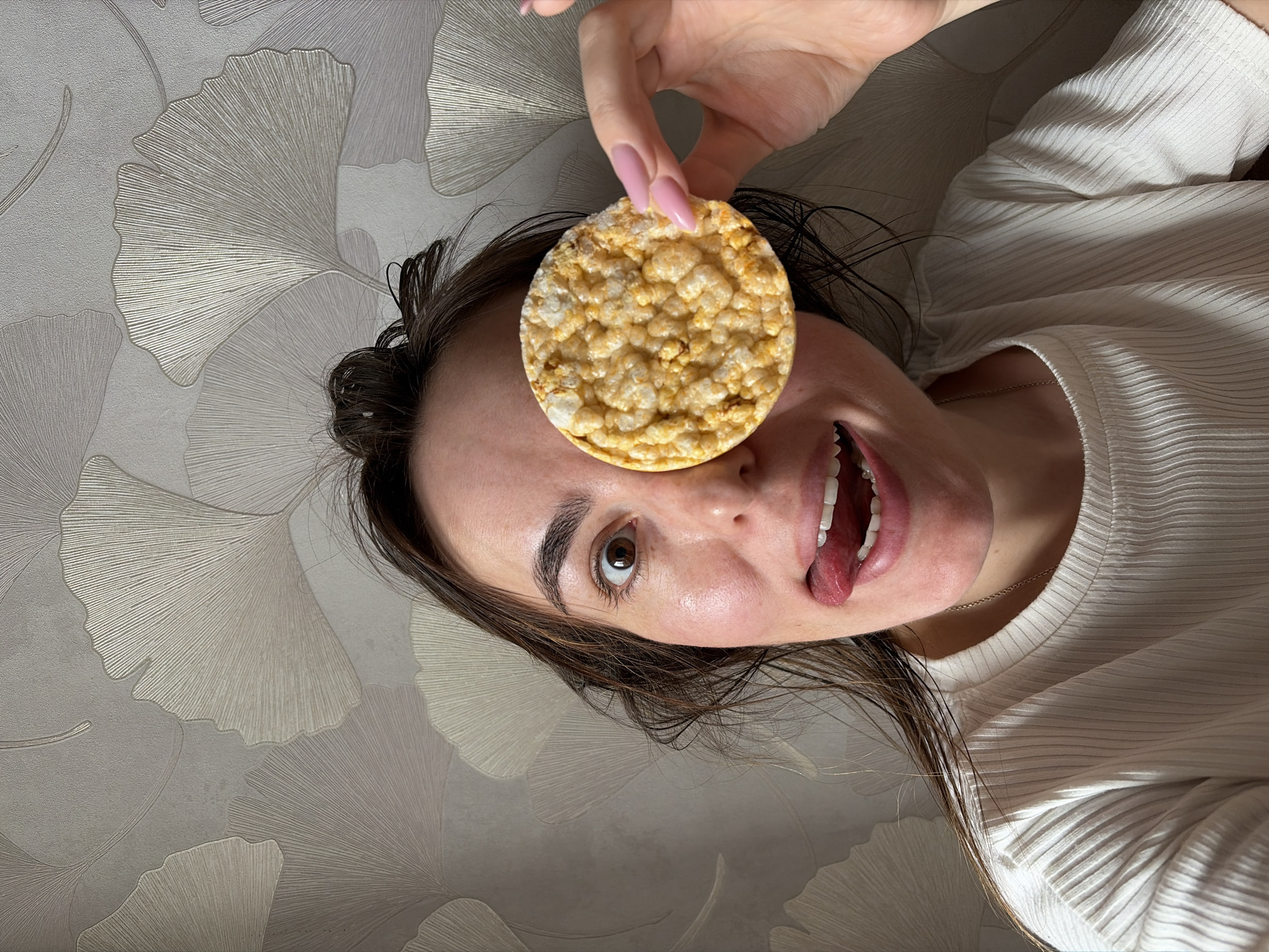 The media file features a woman playfully posing indoors. She is depicted from an overhead perspective, lying down with her head tilted to the side. Her right hand, adorned with long, light pink painted fingernails, holds a circular, golden-yellow puffed snack, resembling a rice or corn cake, directly over her left eye. Her right eye is open and looks towards the viewer. Her mouth is wide open, revealing her teeth, with her tongue playfully sticking out, conveying a humorous or silly emotion. She has dark brown hair spread around her head and is wearing a light-colored, possibly off-white or cream, long-sleeved top with a distinct ribbed texture. A thin silver-colored chain necklace is visible around her neck. The background is a light-toned wallpaper patterned with large, metallic or embossed ginkgo leaves, suggesting an indoor setting. No text is visible in the image.