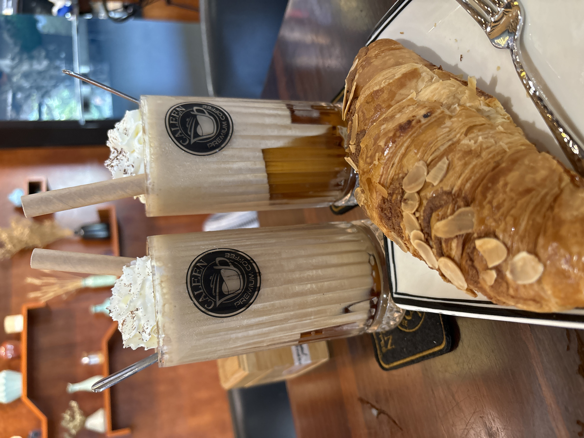 A close-up, eye-level shot captures two tall glasses of iced coffee topped with whipped cream and chocolate shavings, accompanied by an almond croissant on a white plate with a black rim. A silver fork rests beside the croissant. The glasses, featuring a circular black and white logo that reads "CALEEN PREMIUM COFFEE," are filled with a light brown, blended beverage, with visible layers of dark liquid at the bottom. Each glass has a beige paper straw. The food and drinks are arranged on a dark wooden table, with blurred shelves displaying various decorative items in the background, including vases and dried plants. The overall ambiance suggests a cafe setting. The image is professionally lit, highlighting the textures of the croissant and the whipped cream. No people are visible in the frame. The cafe appears to be "CALEEN PREMIUM COFFEE," a place known for its coffee and pastries, likely located in Nha Trang, Vietnam, given the commonality of such establishments in tourist areas there.