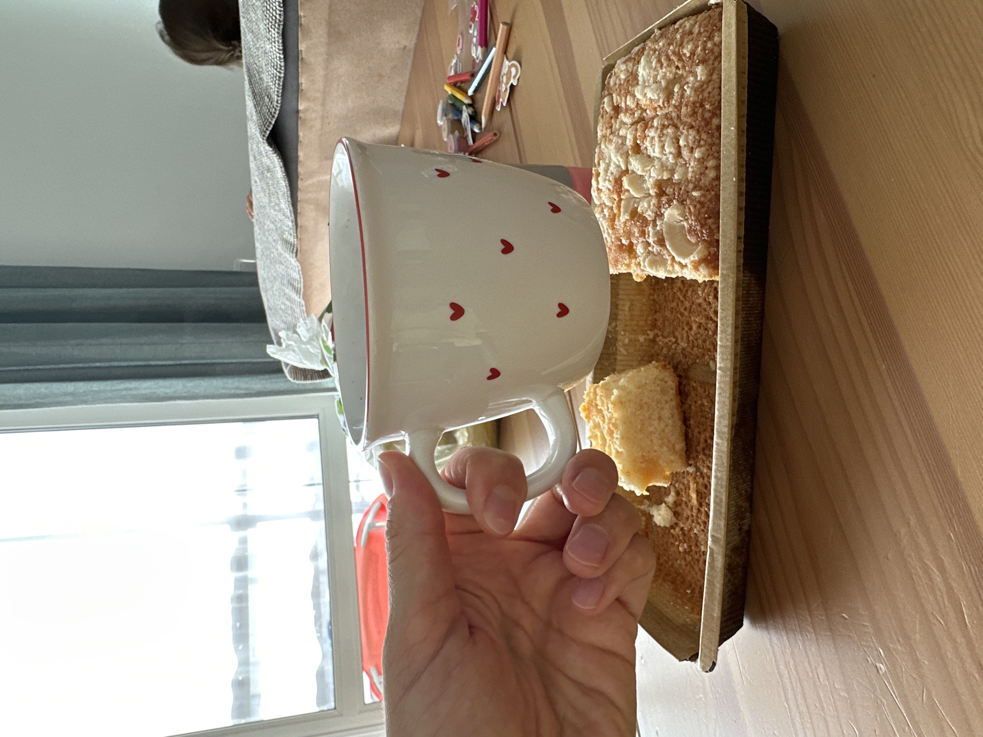 A hand holds a white mug with red hearts on it, positioned over a tray of baked goods. The mug is filled with a light-colored beverage. The tray contains two pieces of cake, one is a square with a crumble topping and the other is a smaller piece. The background shows a window with natural light streaming in, and a blurred area with a person's head visible behind a blanket. There are also art supplies like pencils and markers visible on the table. The scene appears to be indoors, likely in a home.