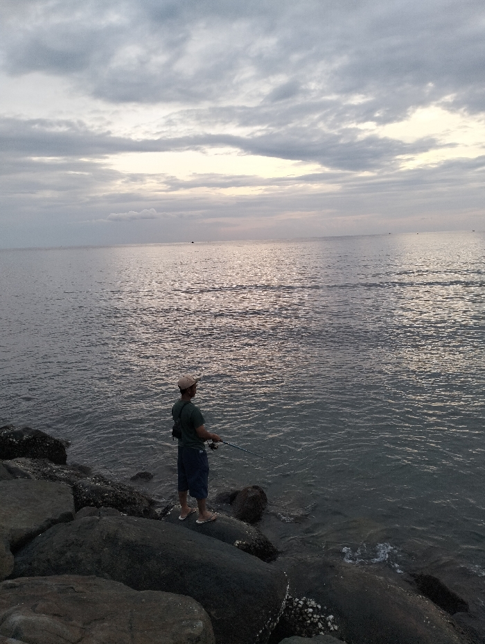 A man fishes from large rocks lining the edge of calm water under a cloudy sky. He wears a hat, dark green shirt, shorts, and sandals. He holds a fishing rod, its line extending into the water. The water is calm with horizontal light reflecting on its surface. The sky is filled with gray and white clouds.