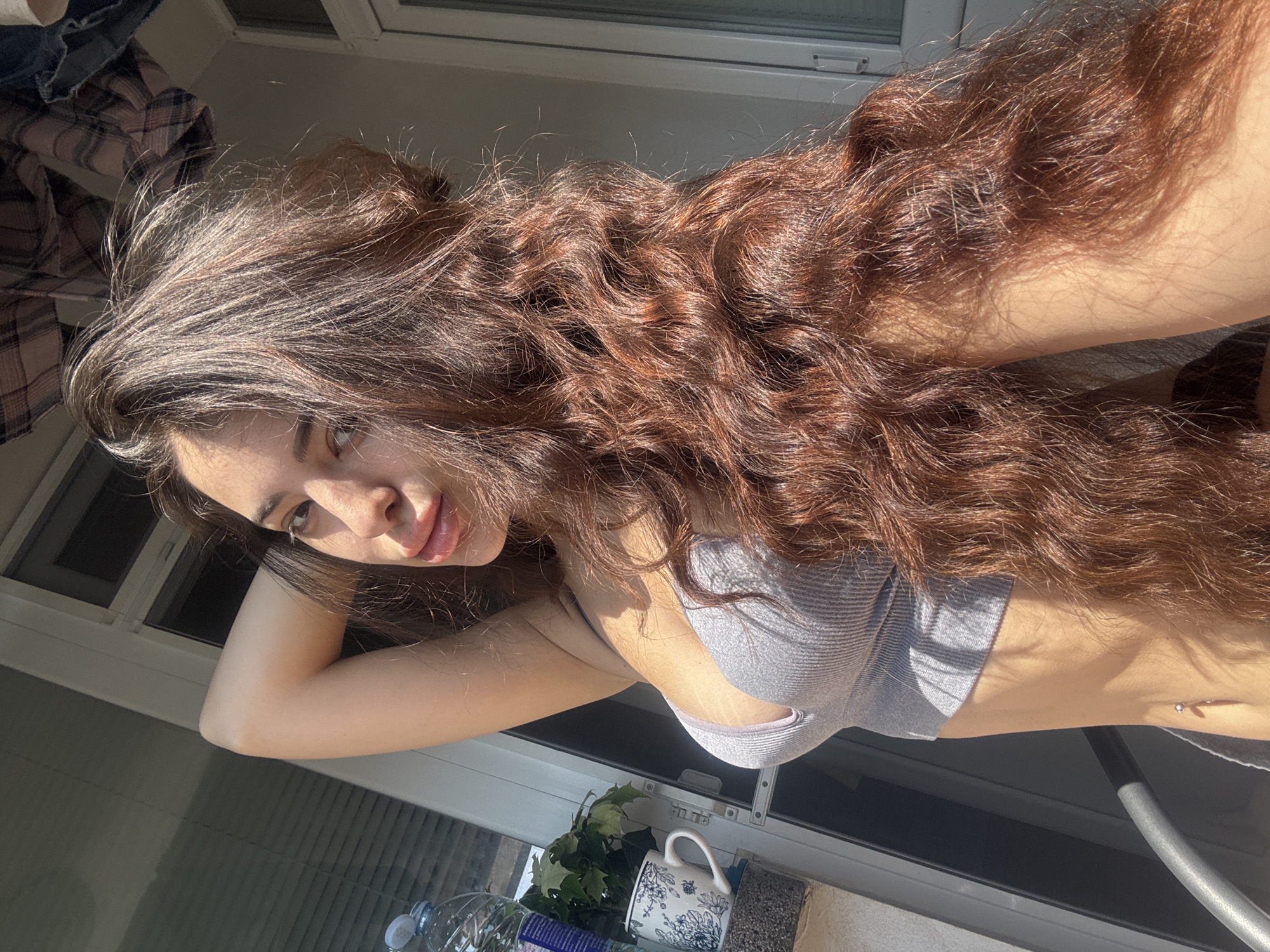 A young woman with long, wavy brown hair is posing in front of a window or possibly on a balcony. She is wearing a grey sports bra and her arm is bent, with her hand near her head. The background shows a windowsill or railing where a white mug with a floral pattern, a plastic bottle, and green leaves are placed. The light suggests it is daytime. The location cannot be confirmed as Plovdiv, Bulgaria based solely on the image provided.