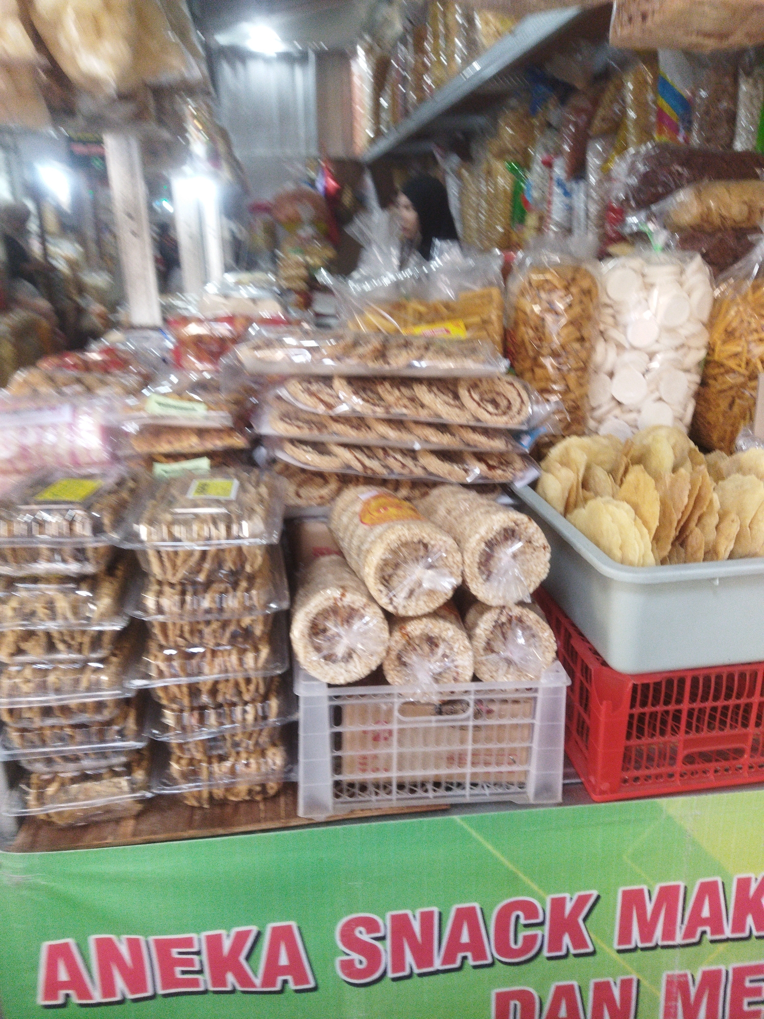 The image depicts a street-side snack stall in Wonogiri, Indonesia. The stall is filled with various packaged snacks, including chips, crackers, and biscuits. In the foreground, a green banner with red text reads "ANEKA SNACK MAK DAN ME". The banner is partially obscured by plastic containers of snacks. The stall appears to be well-stocked, with a variety of items on display. The lighting is bright, suggesting it is daytime. The overall impression is of a bustling market scene.