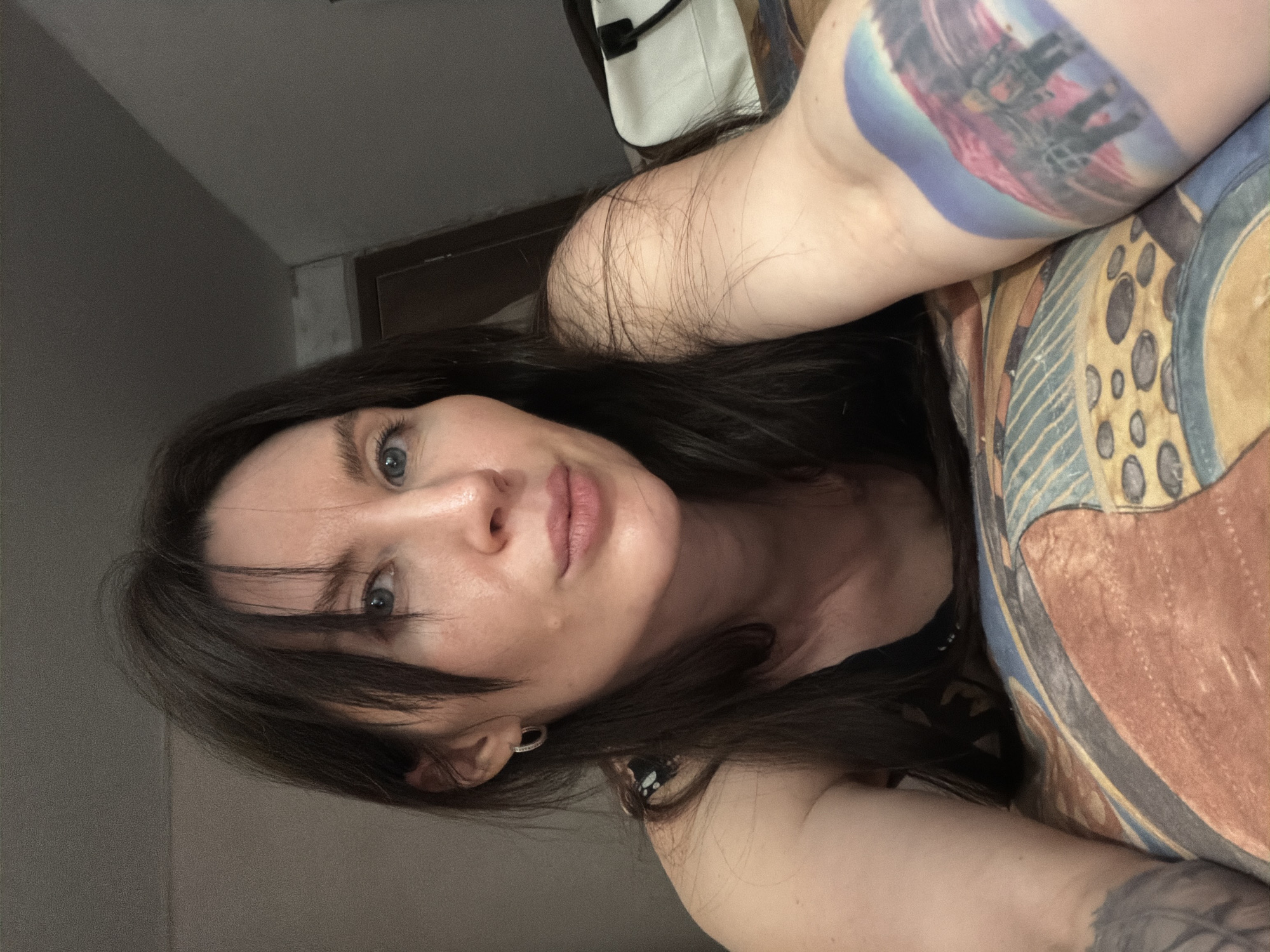 A woman with fair skin, dark hair, and light blue eyes is looking directly at the camera. She has a tattoo on her right upper arm. Her hair falls across her forehead. She is wearing earrings. The background includes a plain wall, a patterned cushion, and a partially visible white bag. There is no indication of the city Perm, Russia.