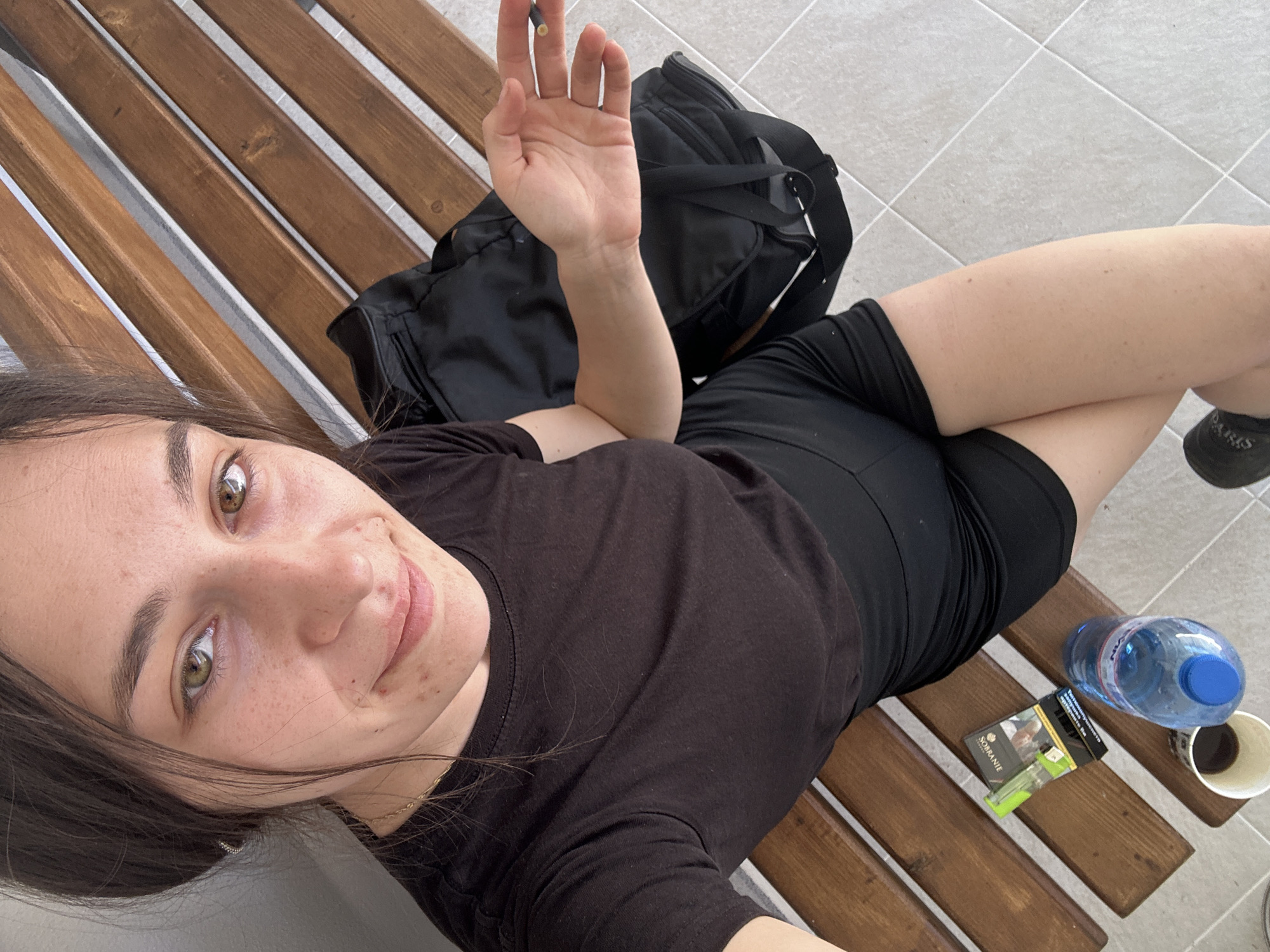 A woman is lying on a wooden bench. She is wearing a black T-shirt and black shorts. Beside her is a black backpack, a bottle of water, cigarettes, and a cup of coffee. She is holding a cigarette in her hand. The background shows a tiled floor. The location is assumed to be in Resen, Bulgaria.