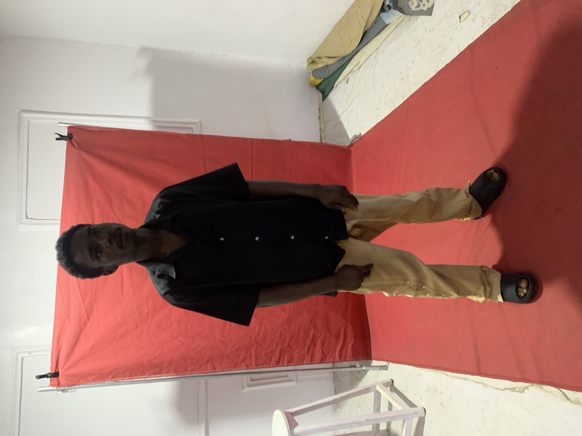 A man stands in a room with a red sheet draped as a backdrop. He wears a black button-down shirt, khaki pants, and dark sandals. The room has white walls, and a white stool sits near the corner. A rolled-up object is present in the upper right corner. The location cannot be definitively determined as Jos, Nigeria, without further evidence.